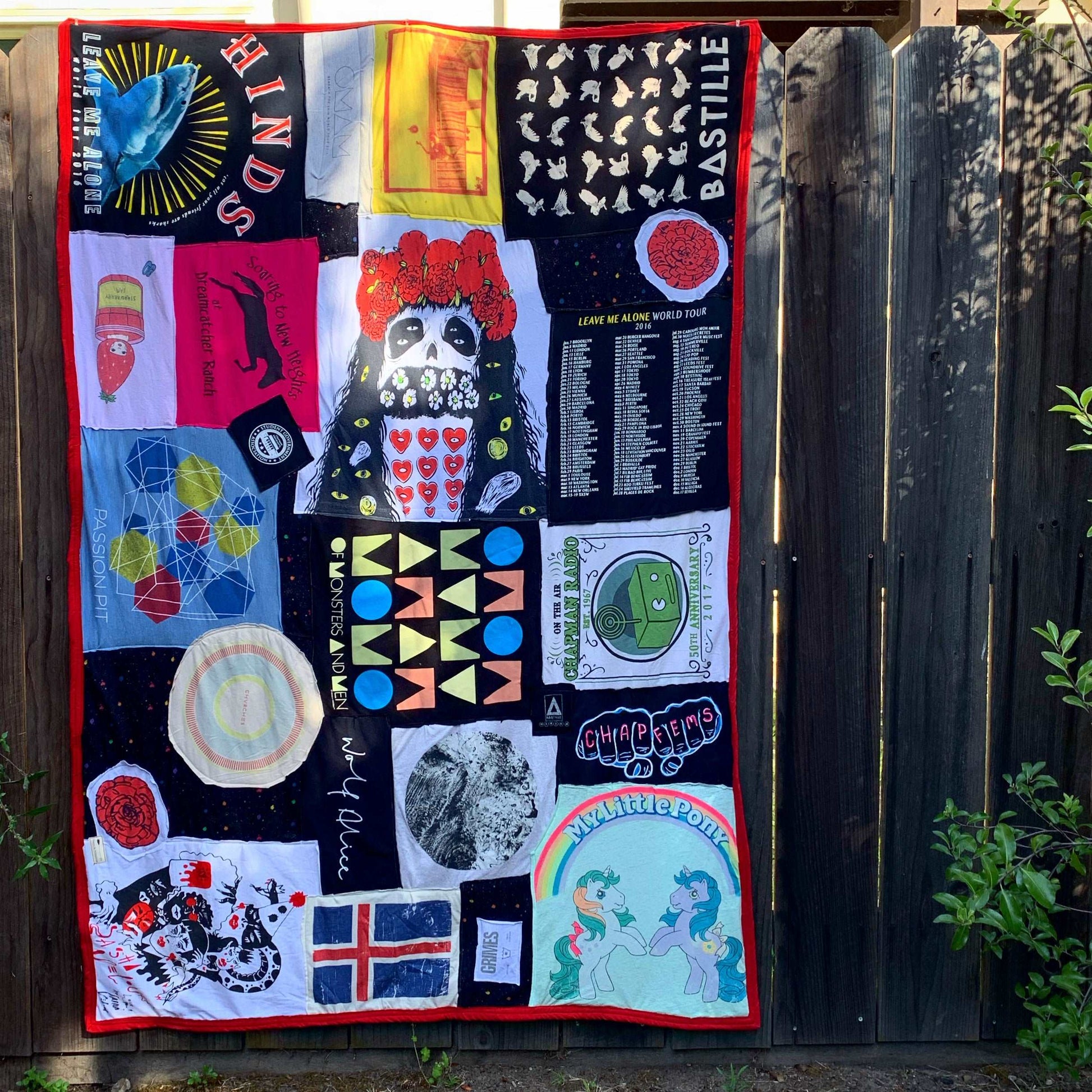 A quilt made fro various t shirts, with a red border, hung up for display on a fence railing.