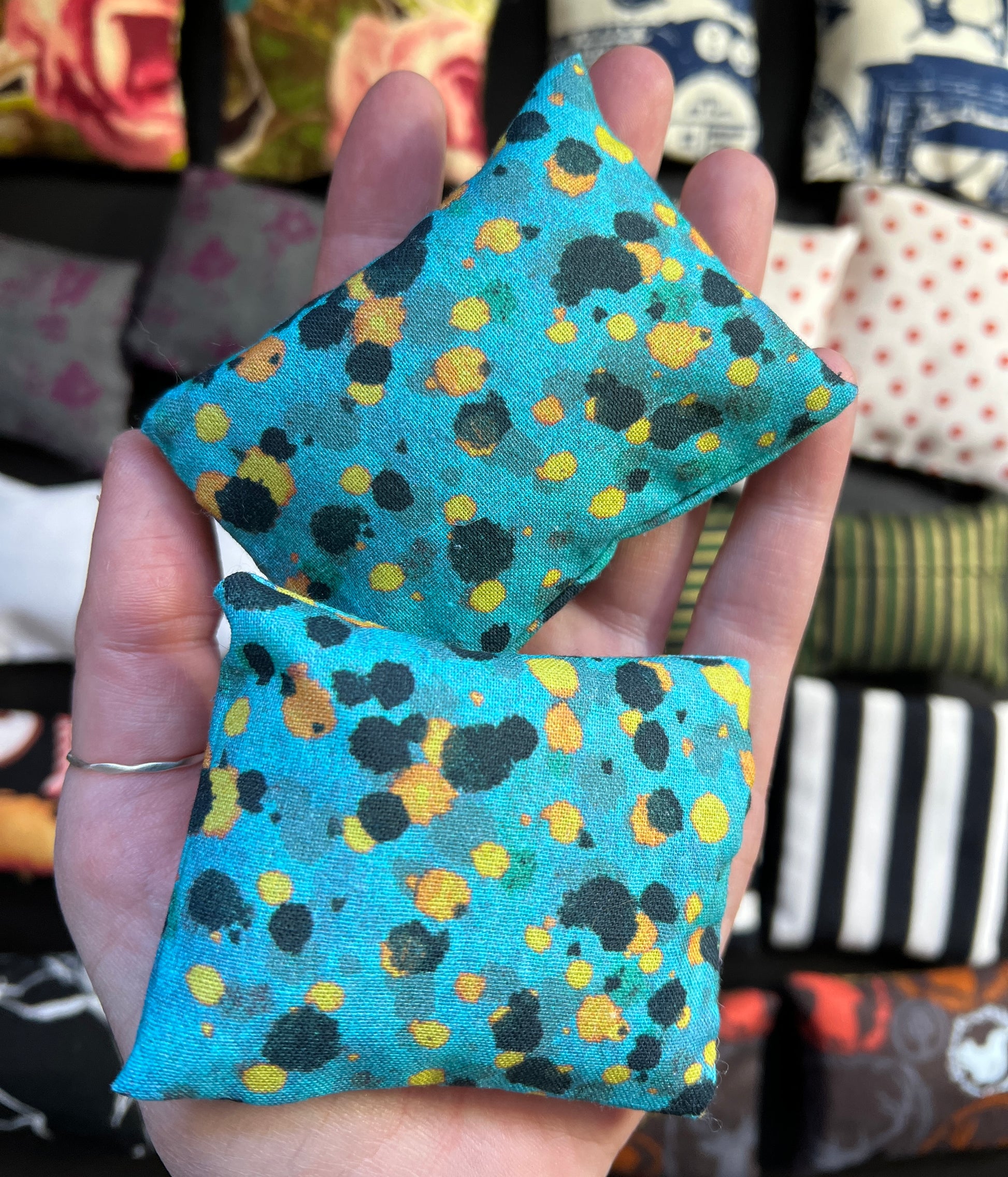 a hand holds a pair of Barbie pillows, for scale. black and yellow paint splotches  on a teal background. other pillows in distance