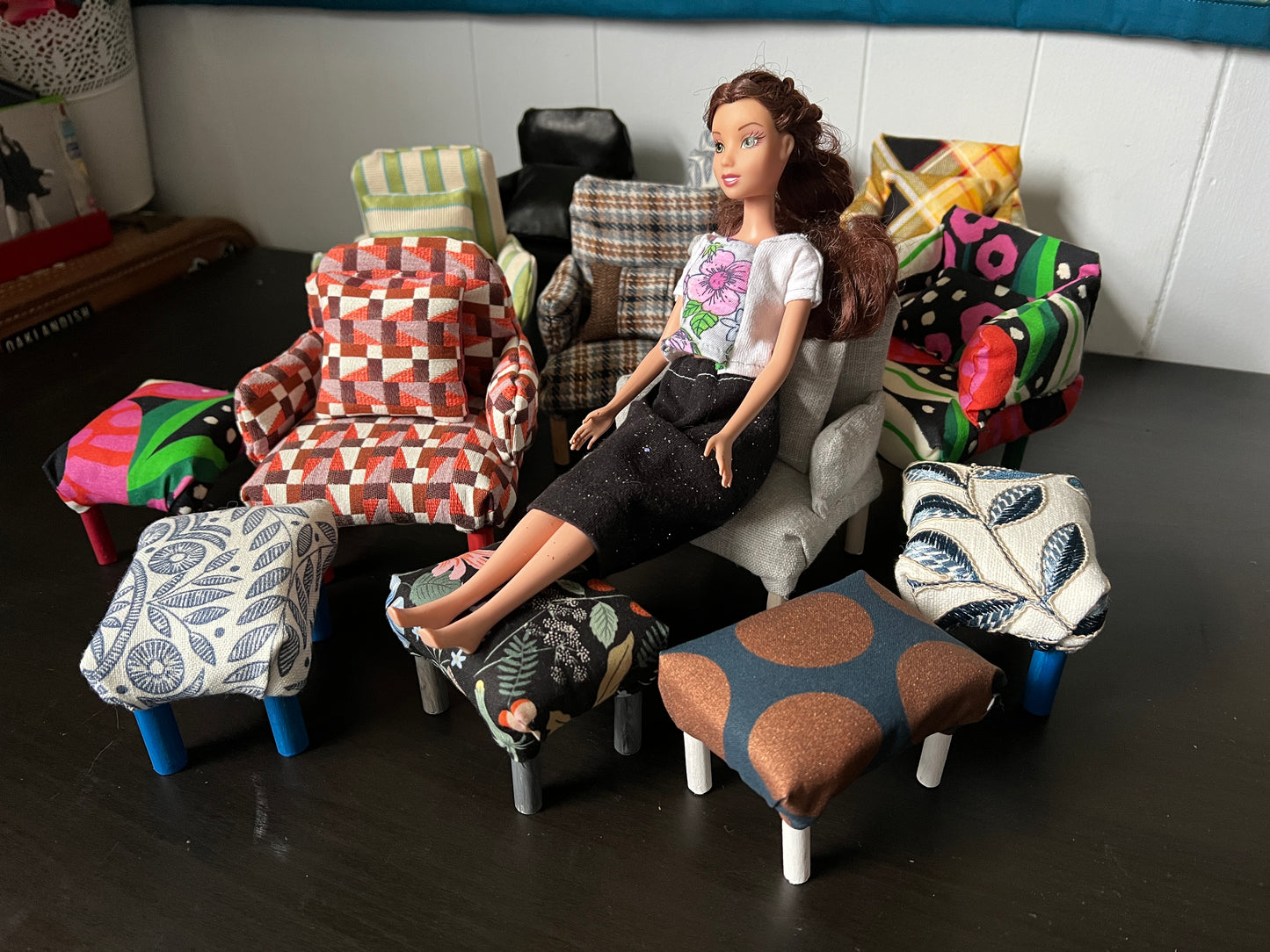 A group of various Barbie-sized dollhouse chairs, and ottomans, barbie is testing out one of the chairs and ottoman