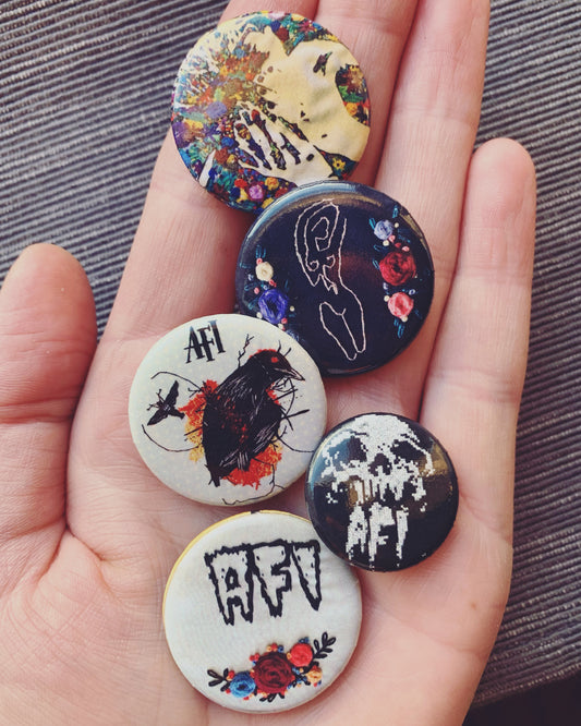 An extended hand holds five pinback buttons featuring various embroidered designs.