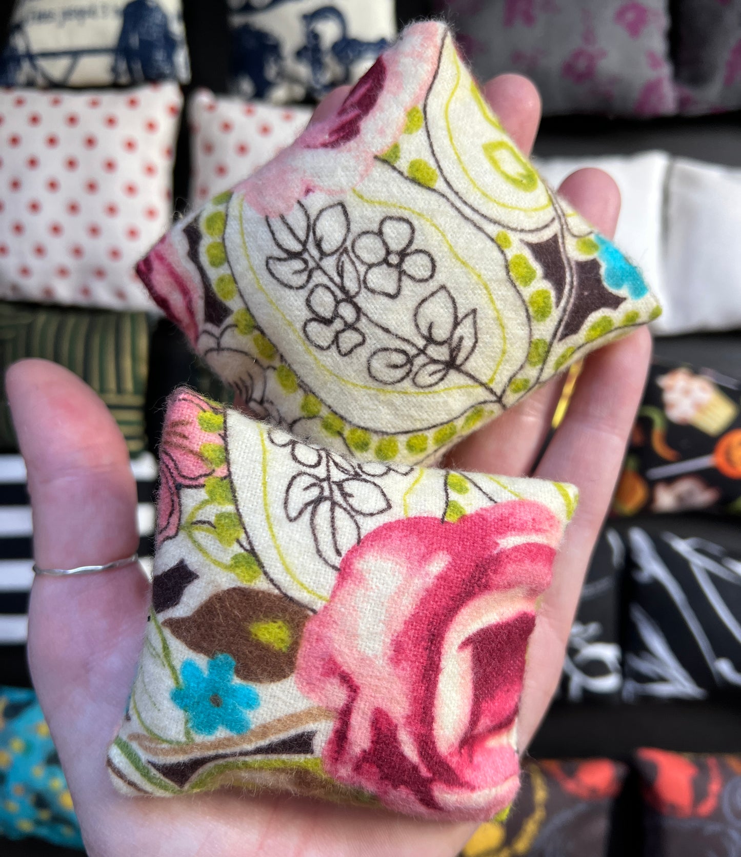 a hand holds a pair of Barbie pillows, for scale. pink rose floral with drawn florals and turquoise flowers. other pillows in distance
