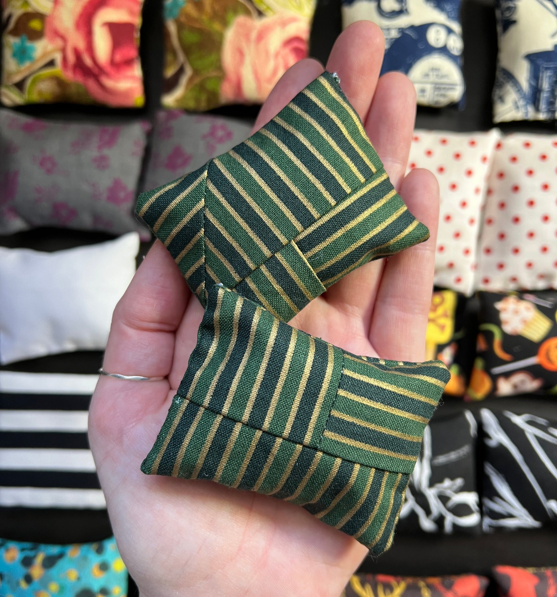a hand holds a pair of Barbie pillows, for scale. dark green, gold, and black stripes, sewn in many directions. other pillows in distance