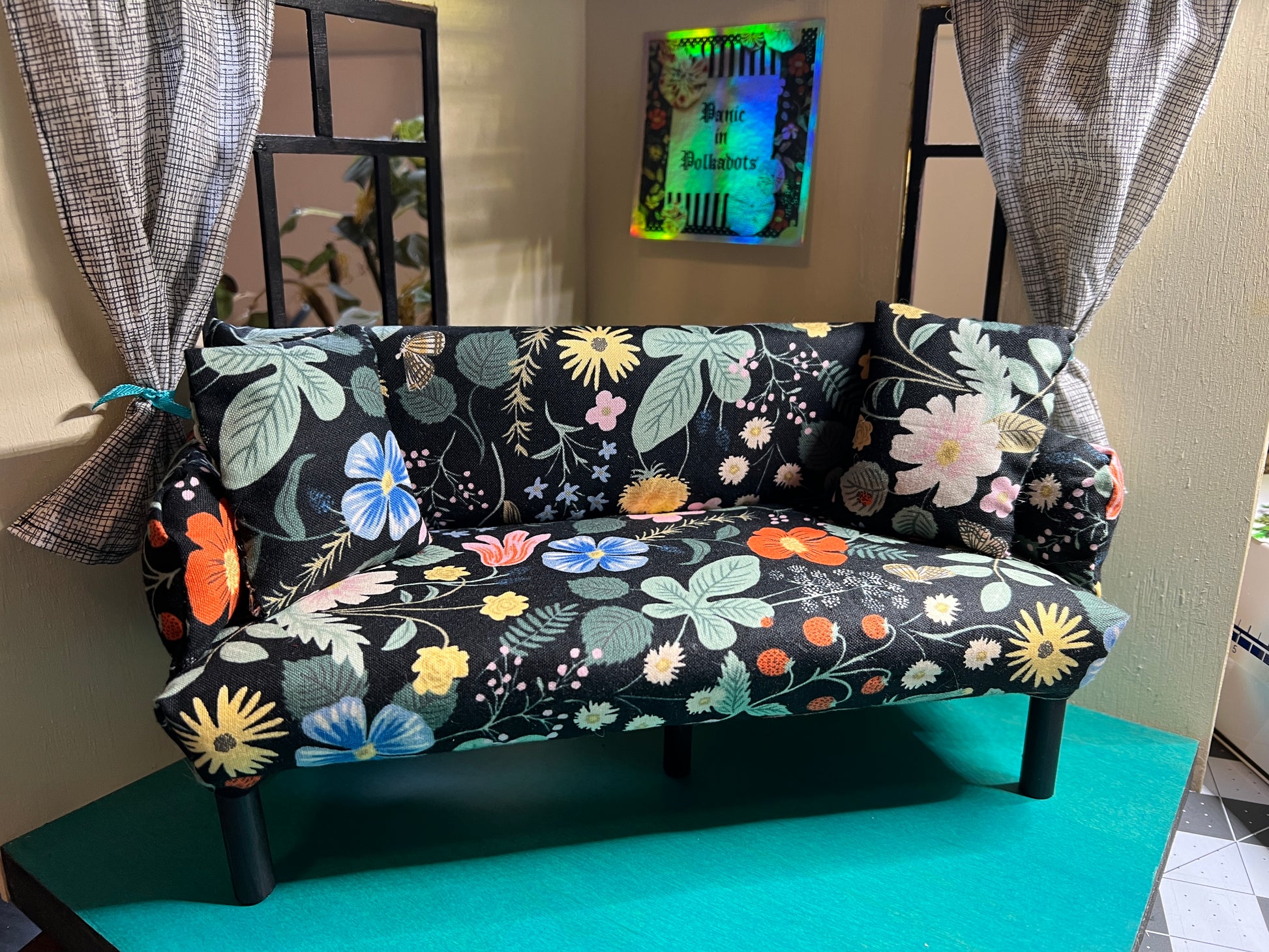 handmade Barbie doll couch, in a dollhouse setting for scale, includes two pillows by Panic in Polkadots. Bold floral fabric in peach, orange, blue, yellow, greenery, and black background