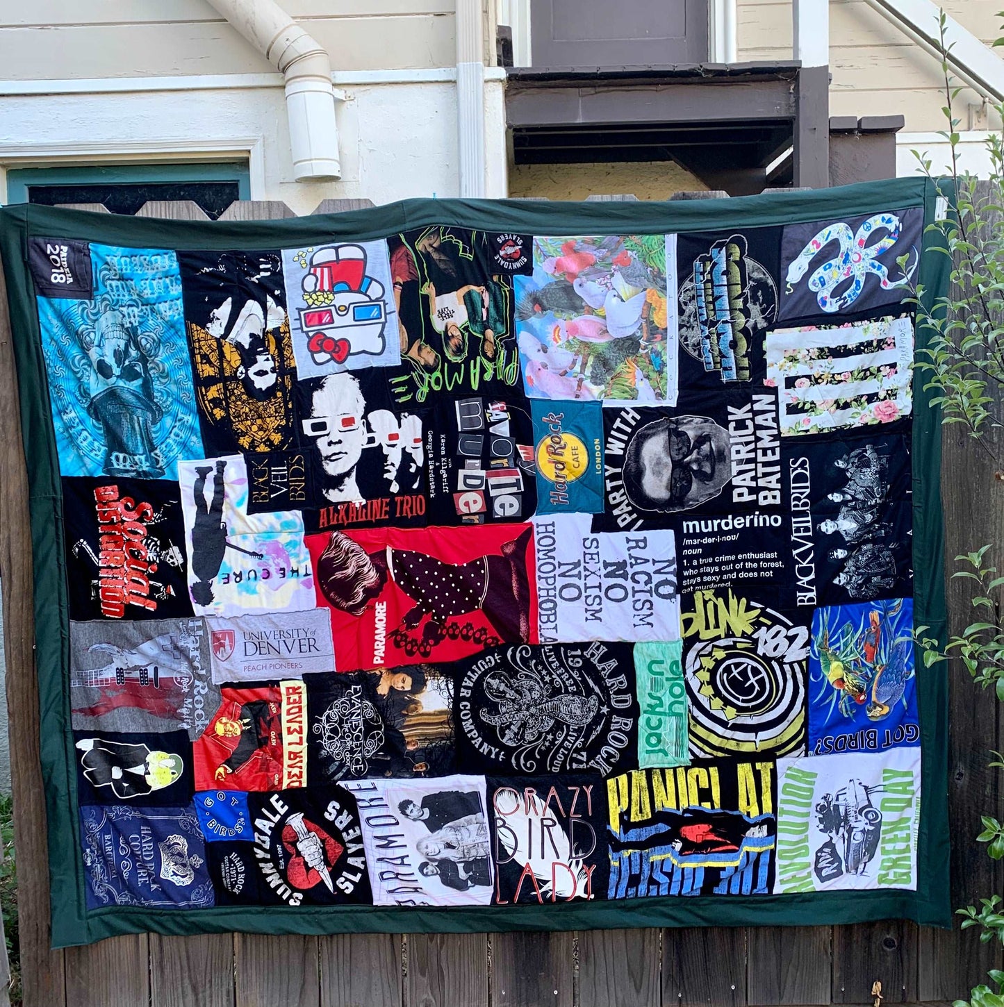 A very large twin sized tshirt quilt, hung up on a fence railing outside, featuring various bands: green day, panic at the disco, social distortion, alkaline trio, the cure, black veil brides 