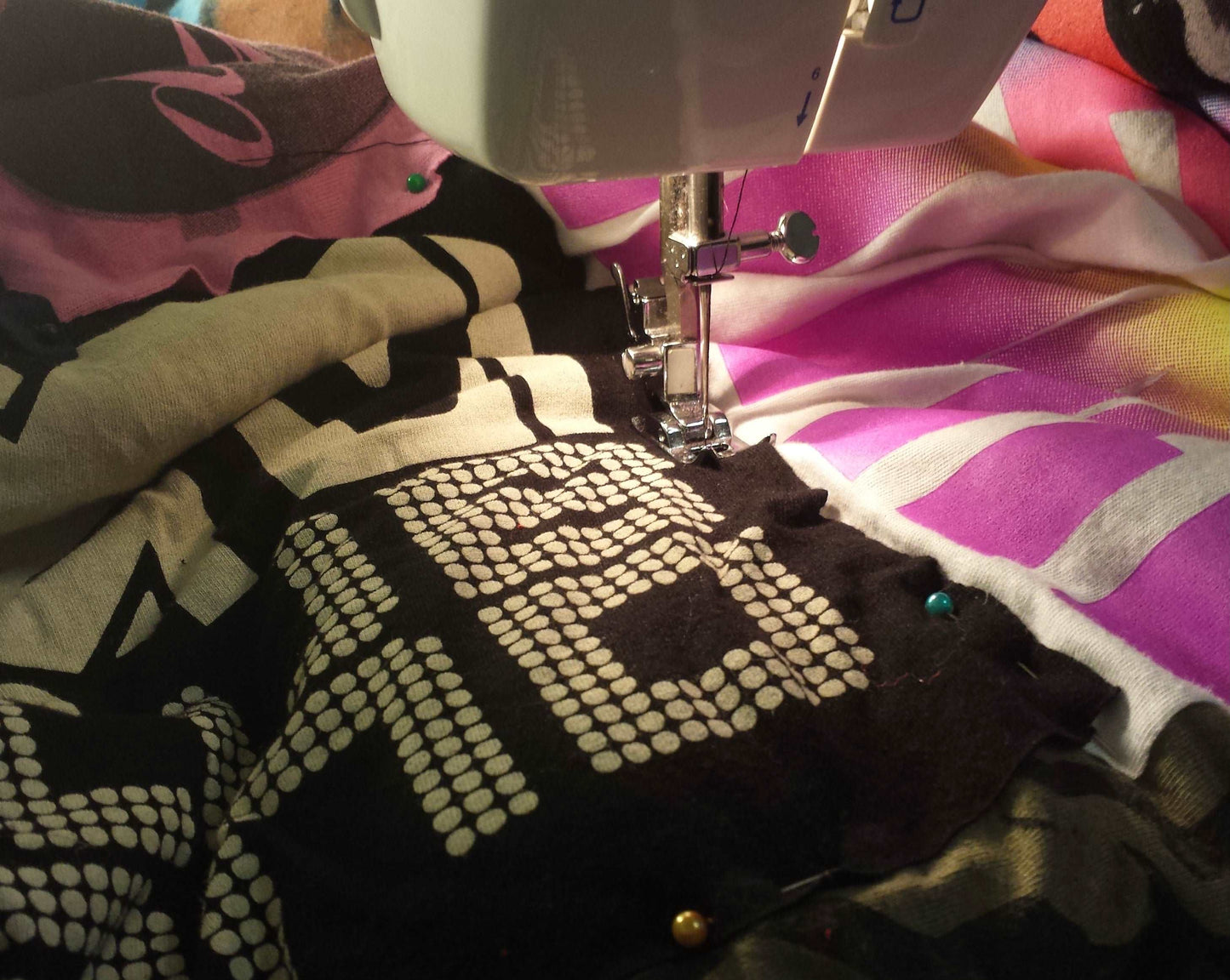 a t shirt quilt, as it is being sewn, under the needle of a sewing machine. there are colorful pins holding the tees in place