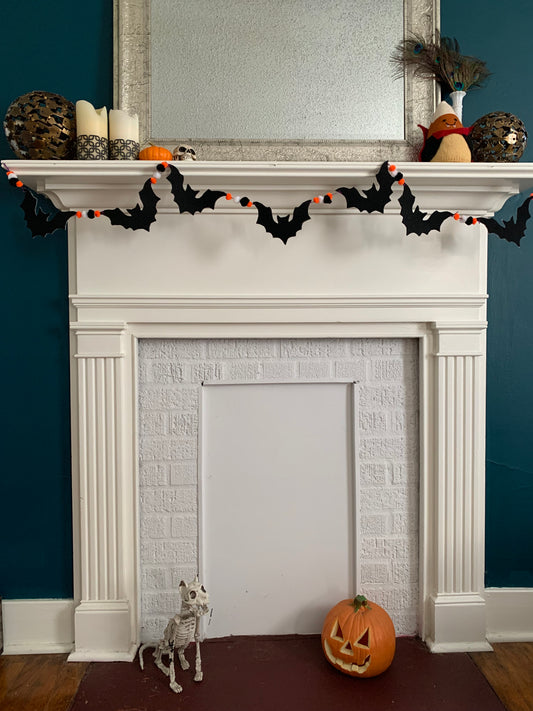a white fireplace with a dark teal wall, adorned with pumpkins and other Halloween decor. A handmade bat garland graces the top of the mantle. 7 felt black bats and various pom poms are strung along the garland.