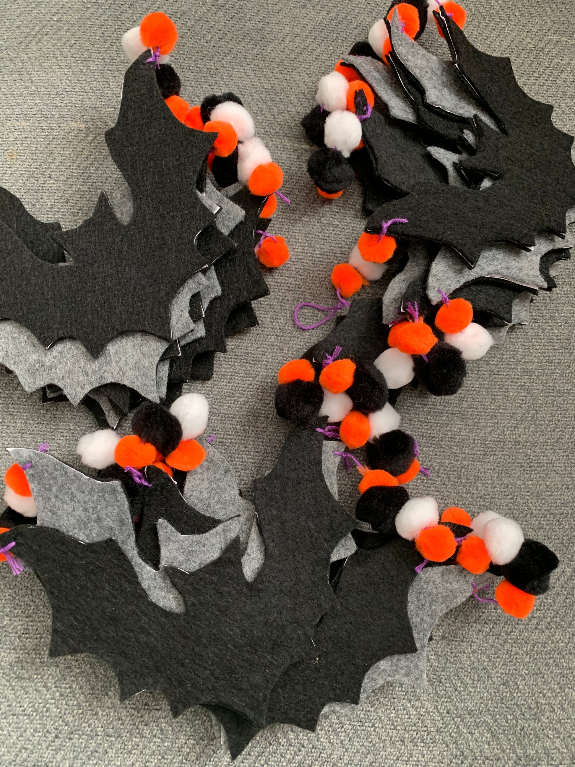 black felt garlands, in a pile, you can see the grey backing of some of the bats. bright orange, white, and black pom poms, and a purple embroidery thread to complete the Halloween look
