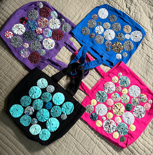a collection of four tote bags, in various colors, with handsewn yoyos on the front of each one. The handles are layered together in the center