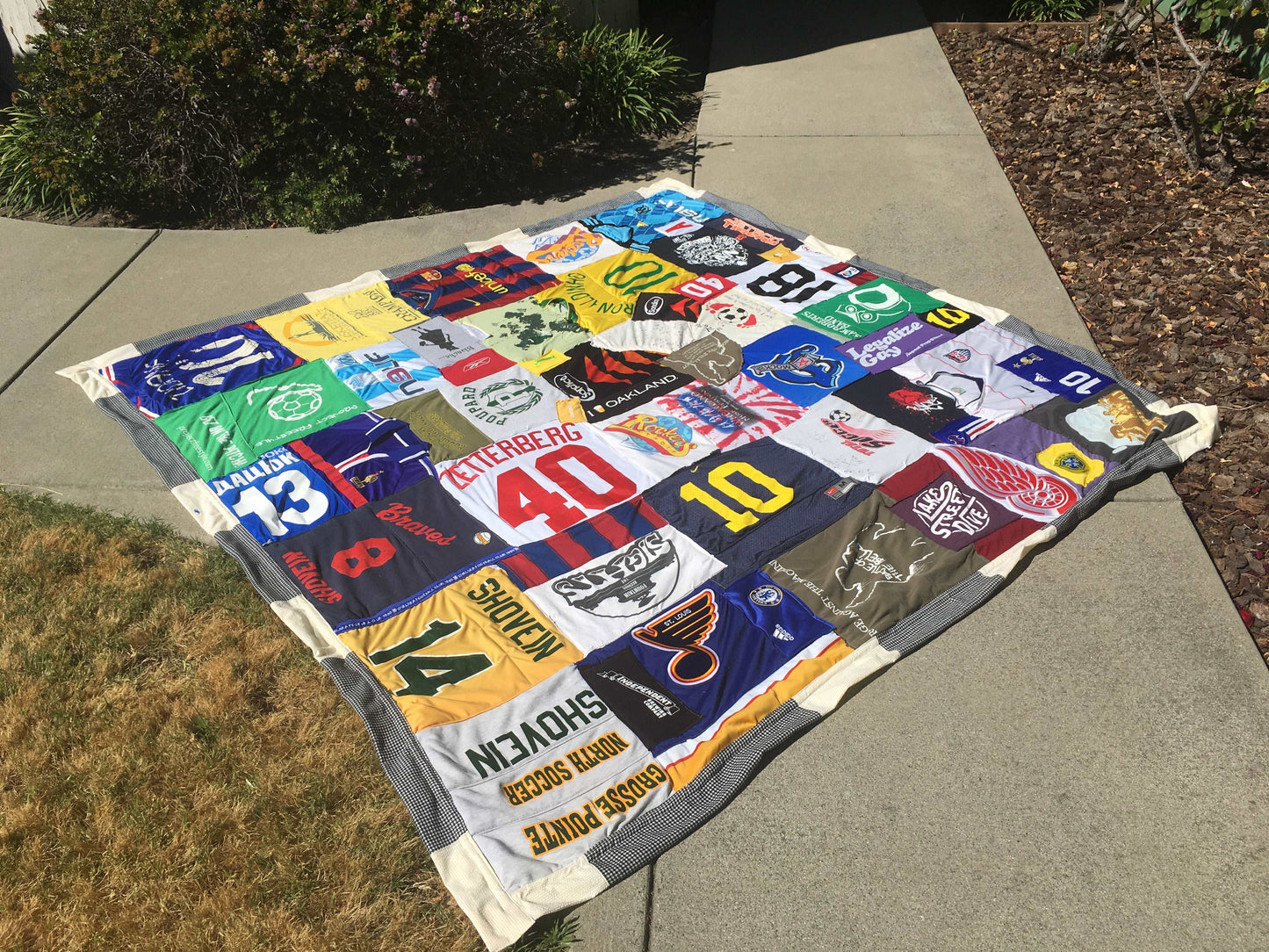 A queen sized quilt created out of Sports Jerseys and tee shirts, is outside on a sidewalk. There are bushes and a lawn in view