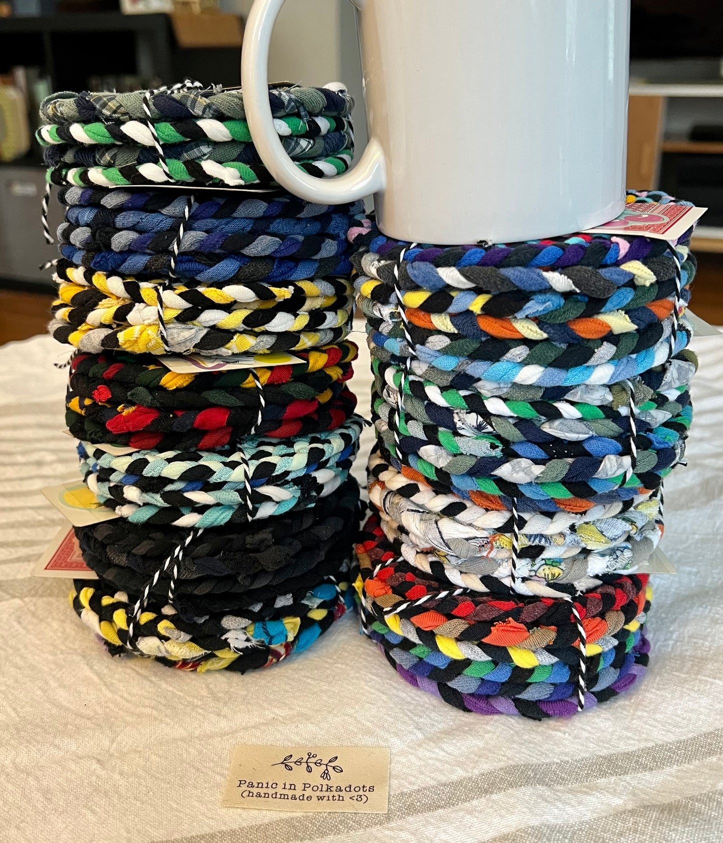 Side view of mini rug coaster bundles, in two stacks, with a white coffee mug on one stack.  Each set is bound by string, with a Panic in Polkadots label in front