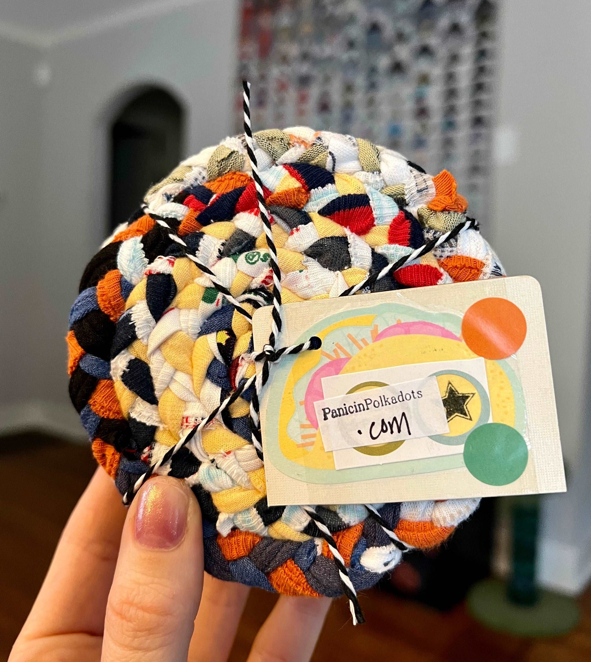 Mini rug coaster set, bound by string and a tag, held up by a hand, with a quilt in the background