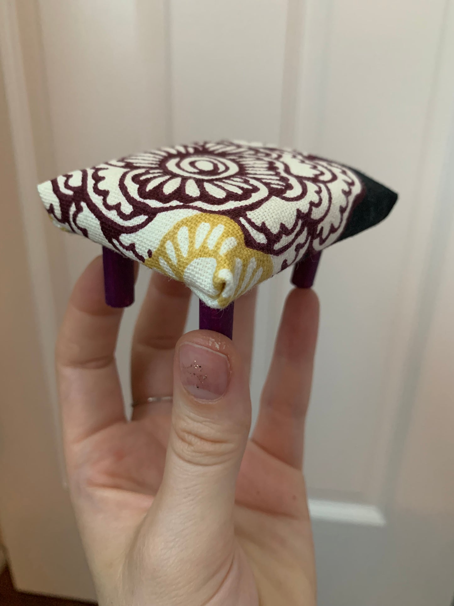 a miniature dollhouse ottoman, held in a hand for scale. This one is white with floral design, and dark purple legs