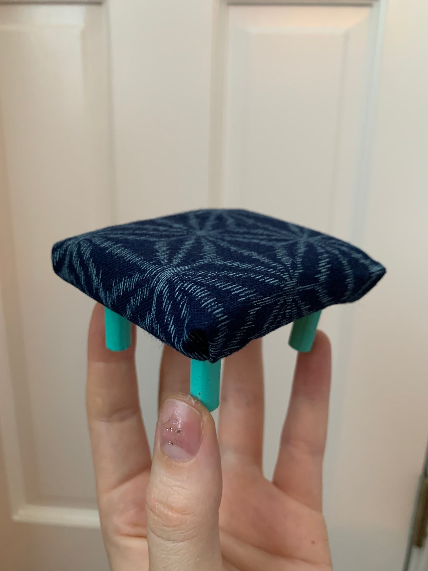 a miniature dollhouse ottoman, held in a hand for scale. This one is navy with a geometric design and turquoise legs