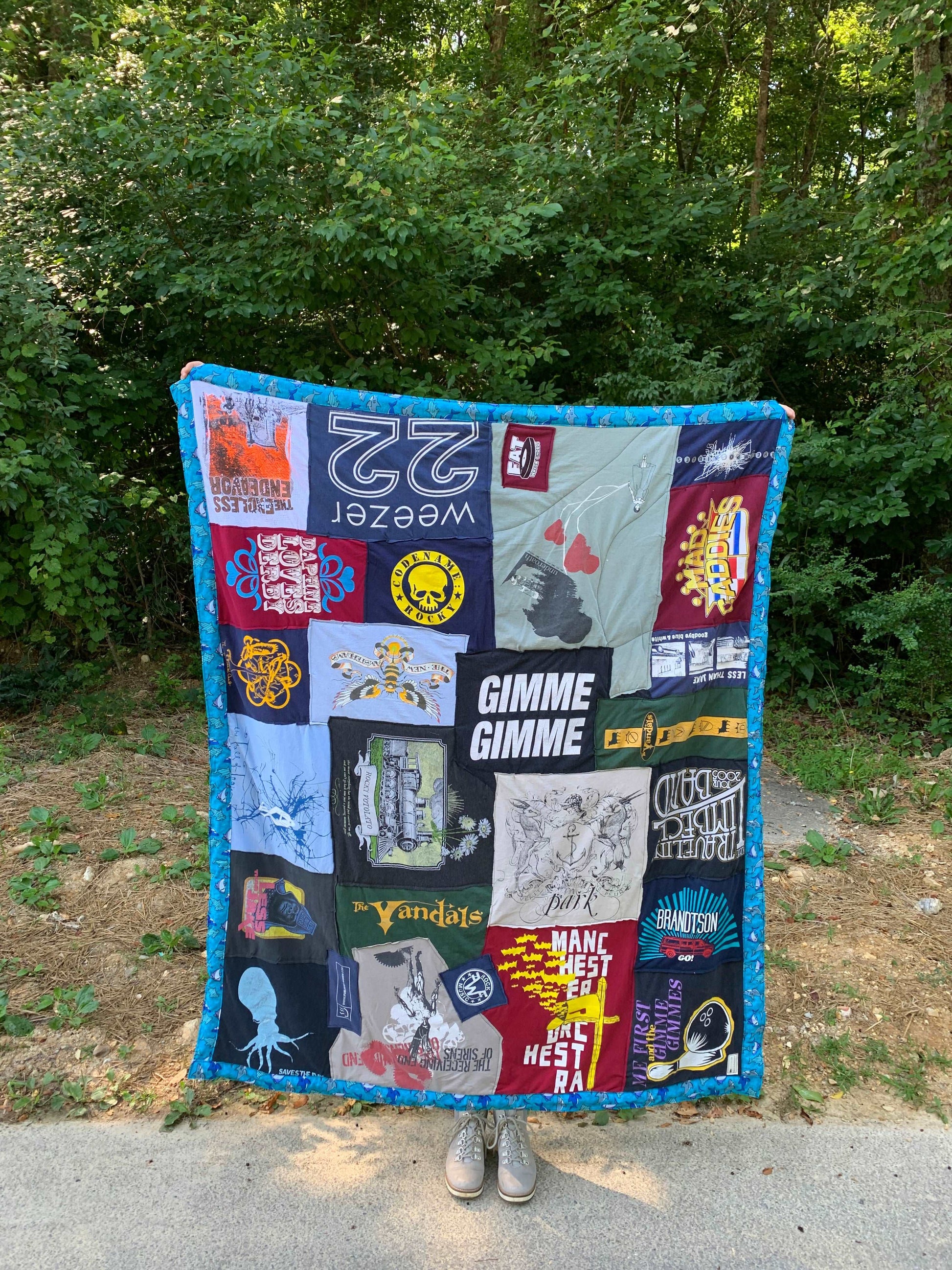 A tshirt quilt is held up outside on a sidewalk, and it have bad tees from: weezer, the Vandals, Me First and the Gimme Gimmes , with a blue shark border