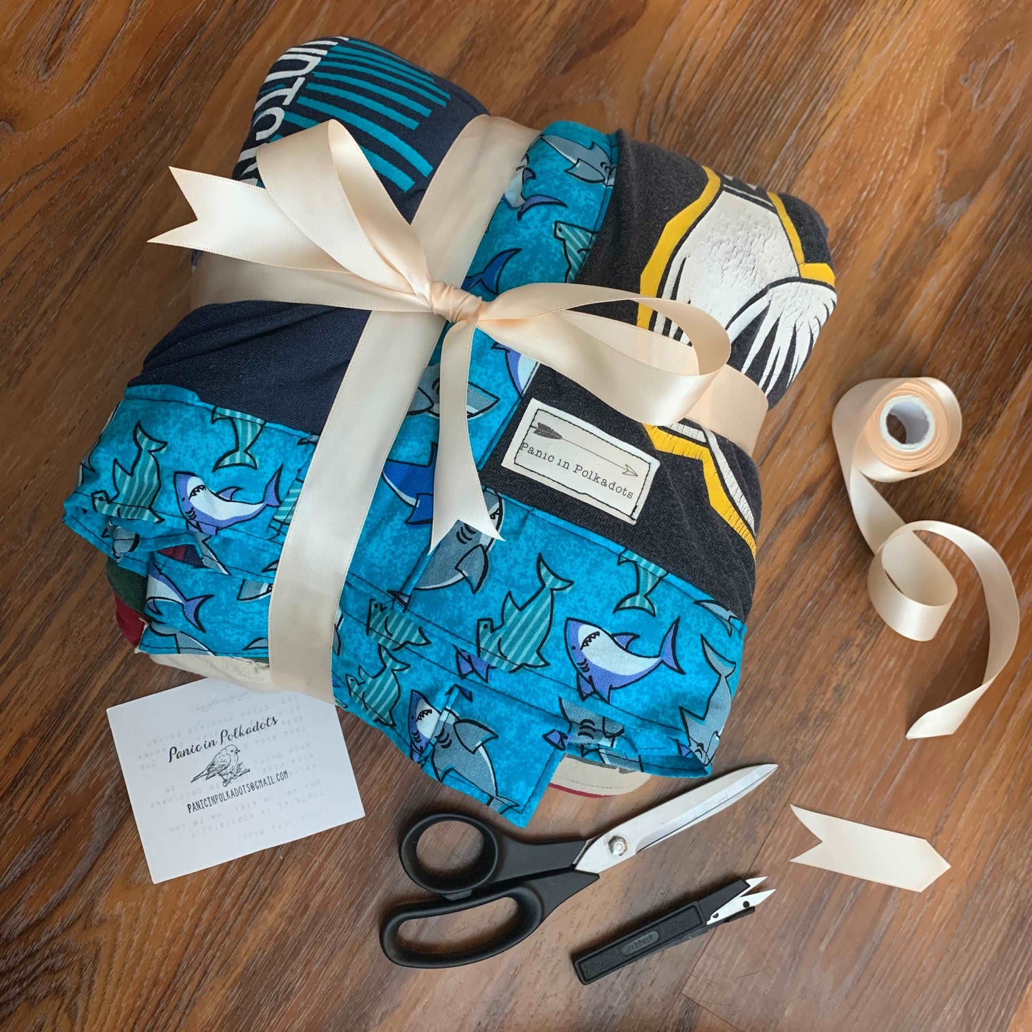 a custom tshirt quilt, with blue shark border, all folded up and wrapped up with a ribbon. A pair of gingham scissors, and snips, and a card with Panic in Polkadots, are in view