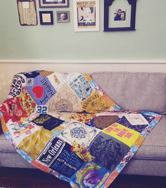 A colorful custom quilt, draped over a grey couch, with a few picture frames on the wall above