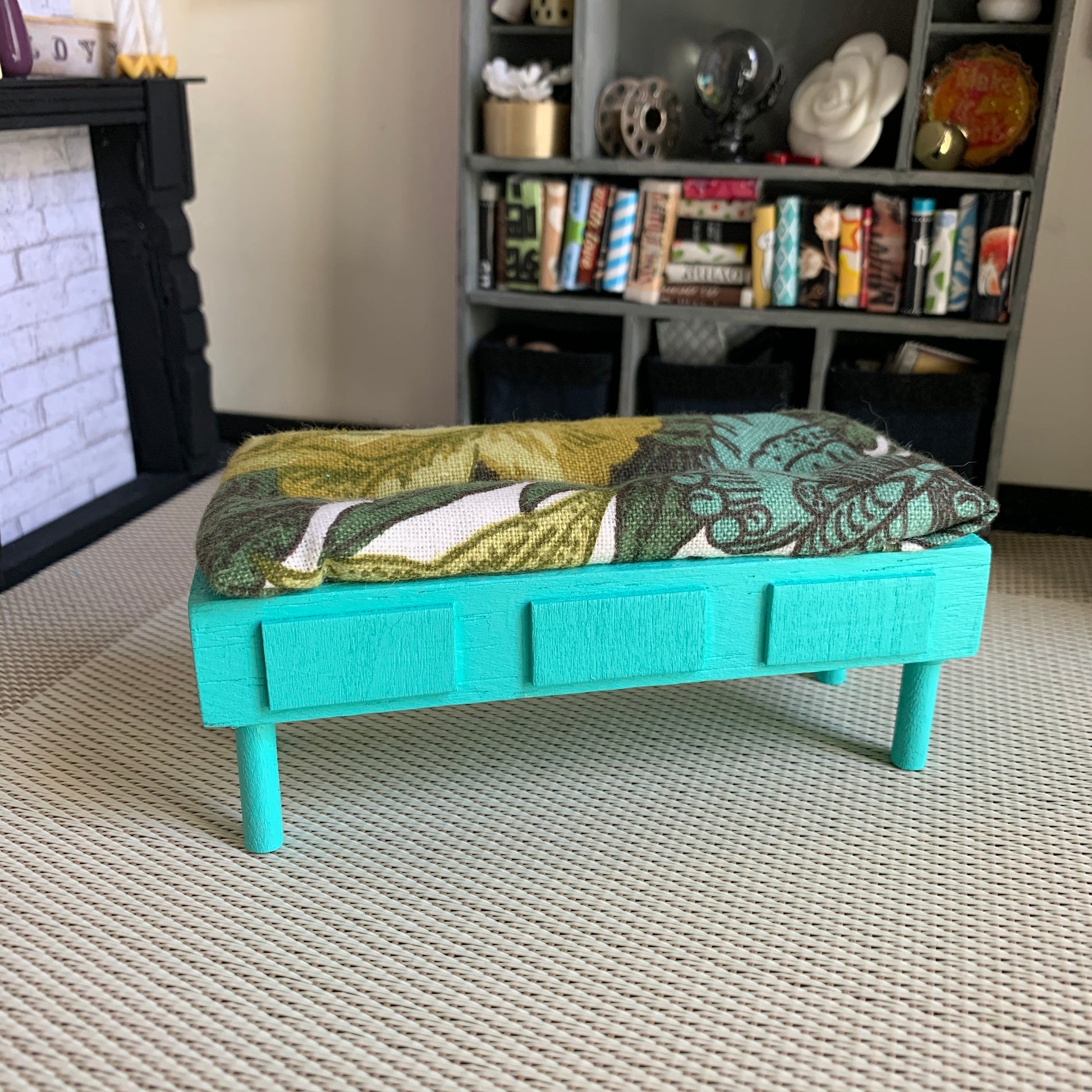 a miniature dollhouse bench, in a dollhouse room for scale. This one has a green and teal floral cushion, with turquoise "storage" and legs