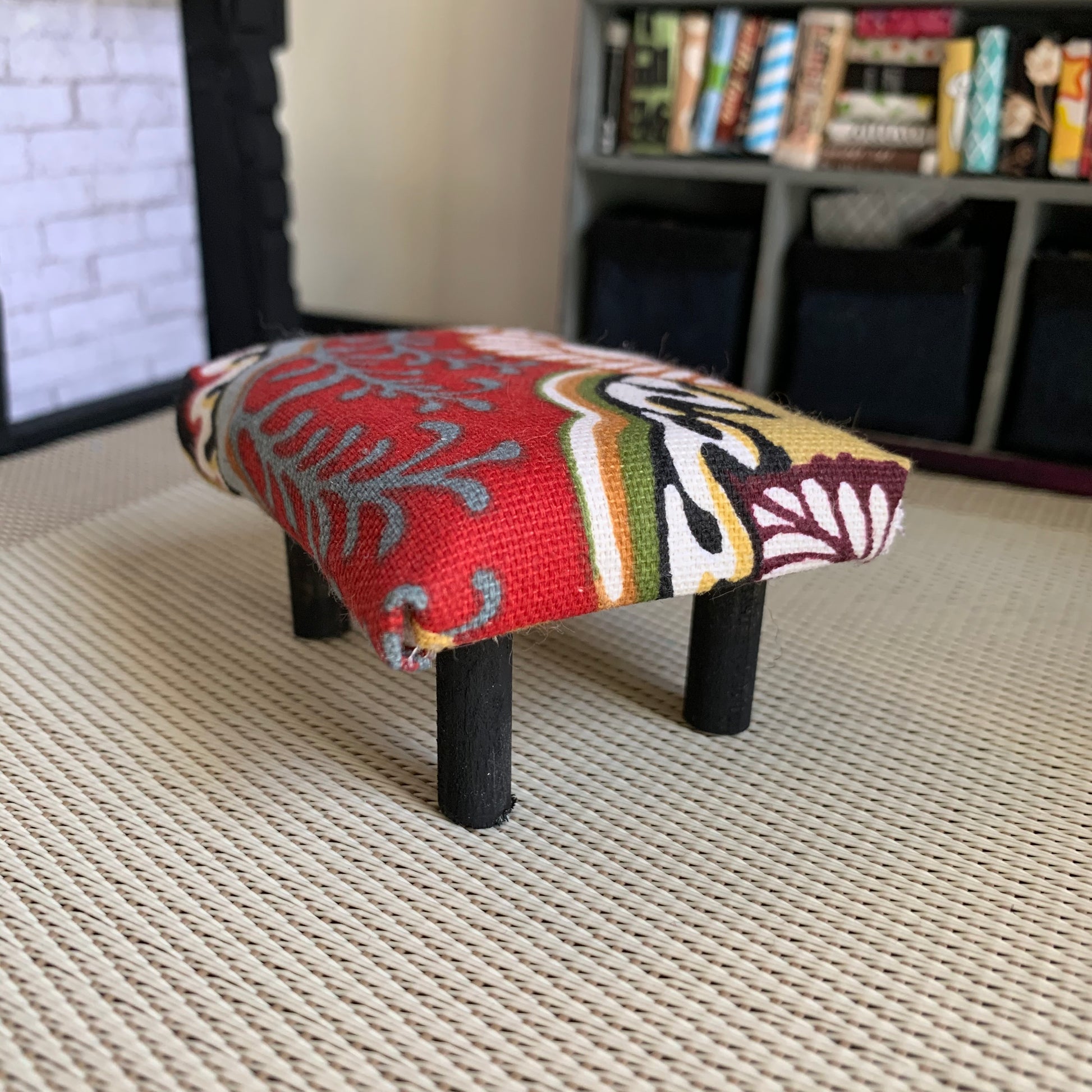 a miniature dollhouse ottoman, in a dollhouse room for scale. This one is red with white and grey floral, with black legs