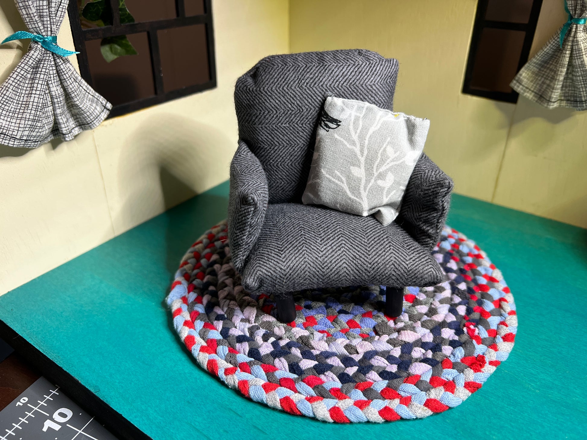 a miniature handmade dollhouse chair, on top of a mini rug (sold seperately) in a dollhouse room box. This one is a dark grey herringbone with light grey floral accent pillow