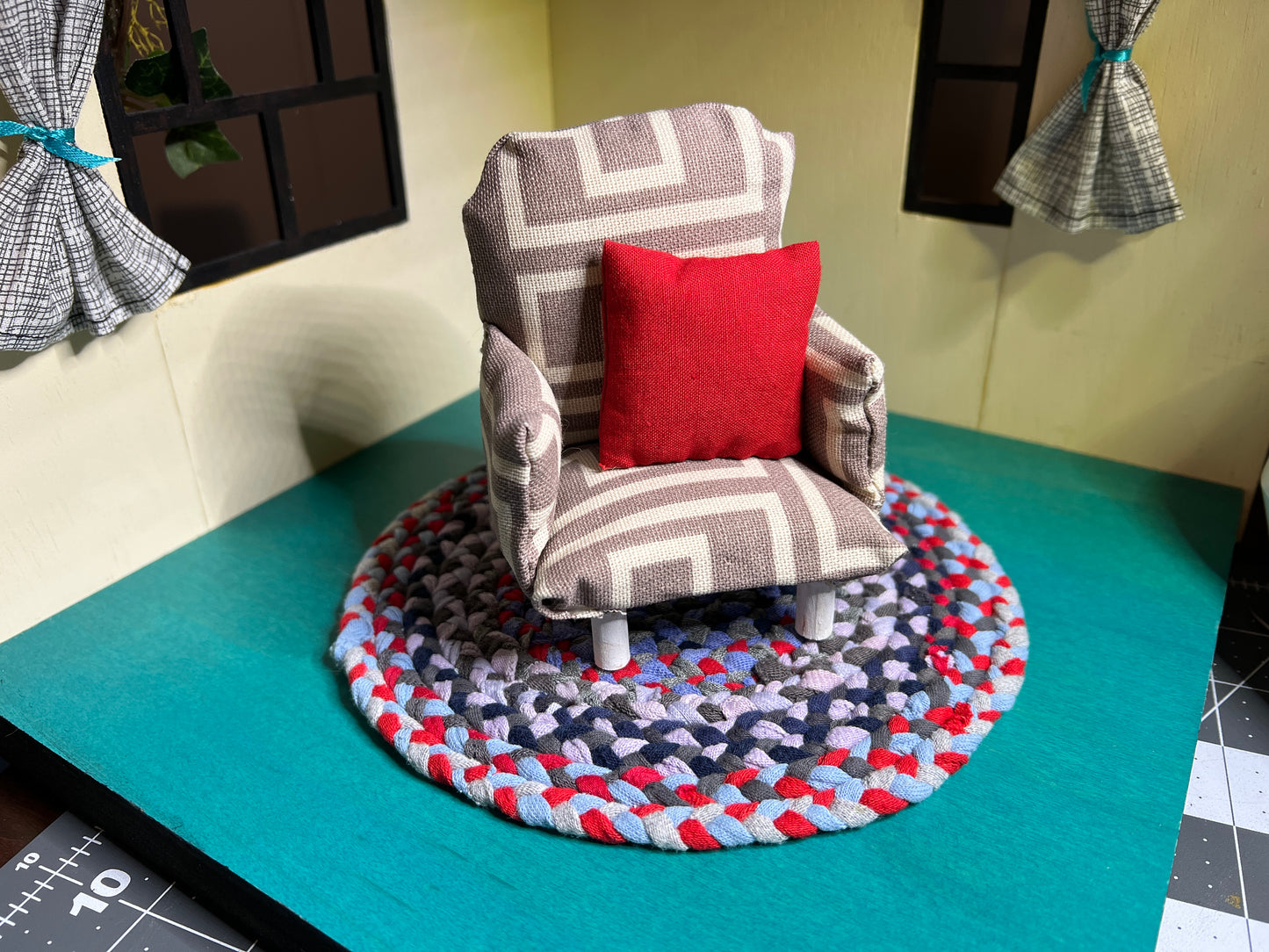 a miniature handmade dollhouse chair, on top of a mini rug (sold seperately) in a dollhouse room box. This is a grey geometric chair with a red accent pillow