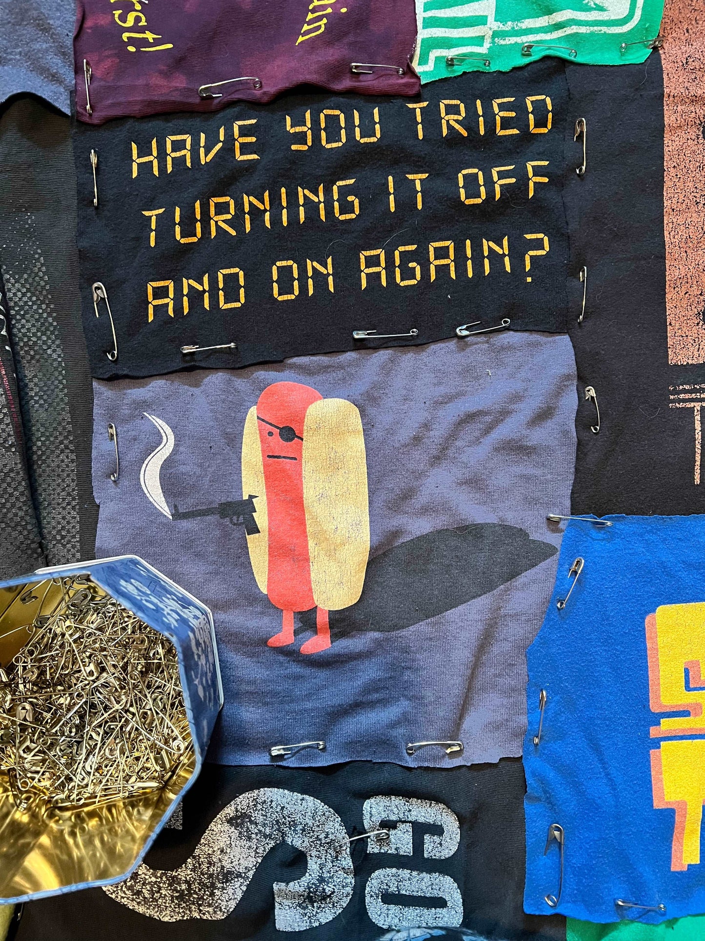 closeup of a tshirt quilt, all pinned and ready to sew. A hotdog with an eye patch holds a smoking gun, above it are the words "Have you tried turning it off and on again"