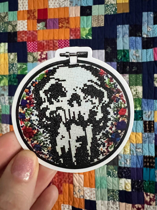 AFI skull sticker in cross stitch with embroidered flower detail around edges, embroidery hoop in shaped sticker. held up against a colorful background