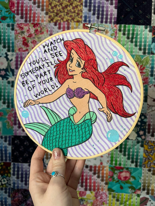 Ariel from The Little Mermaid, on an embroidery hoop, held up against a colorful background. words "Watch and You'll see, someday I'll be, part of your world"