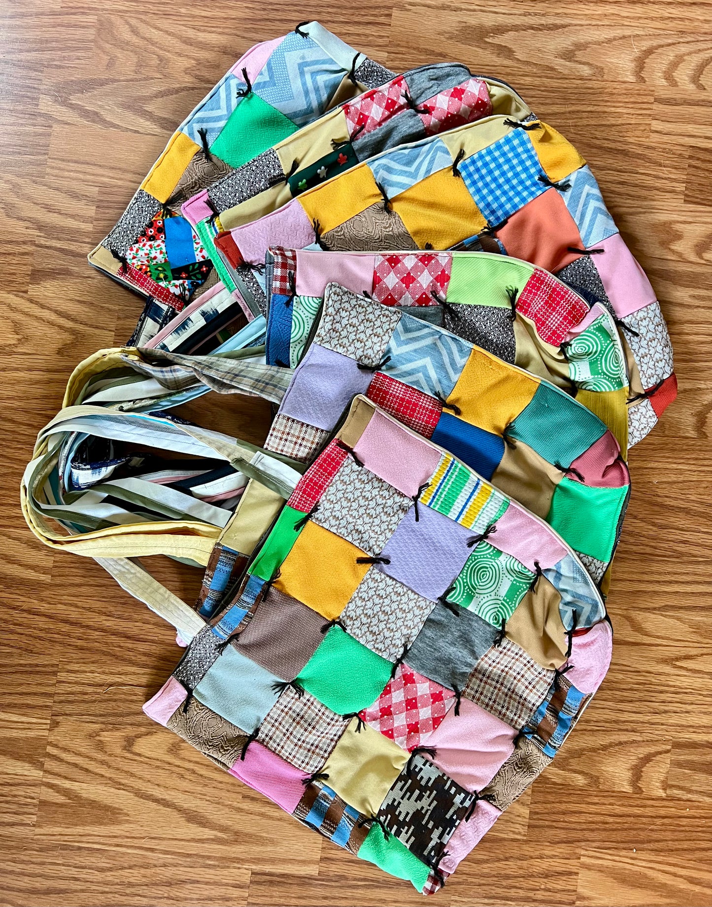 A handmade patchwork fabric tote bag with a multicolor design, laid flat on a wooden floor. The bag features various patchwork squares in pink, yellow, blue, green, and brown, with a striped fabric shoulder strap.