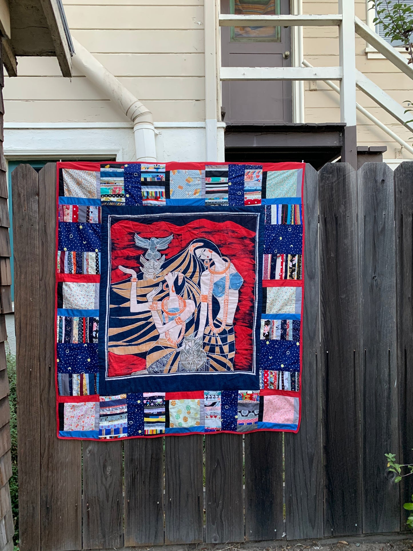 the wall hanging quilt is on display outside on a fence railing, this is a little farther away of a shot