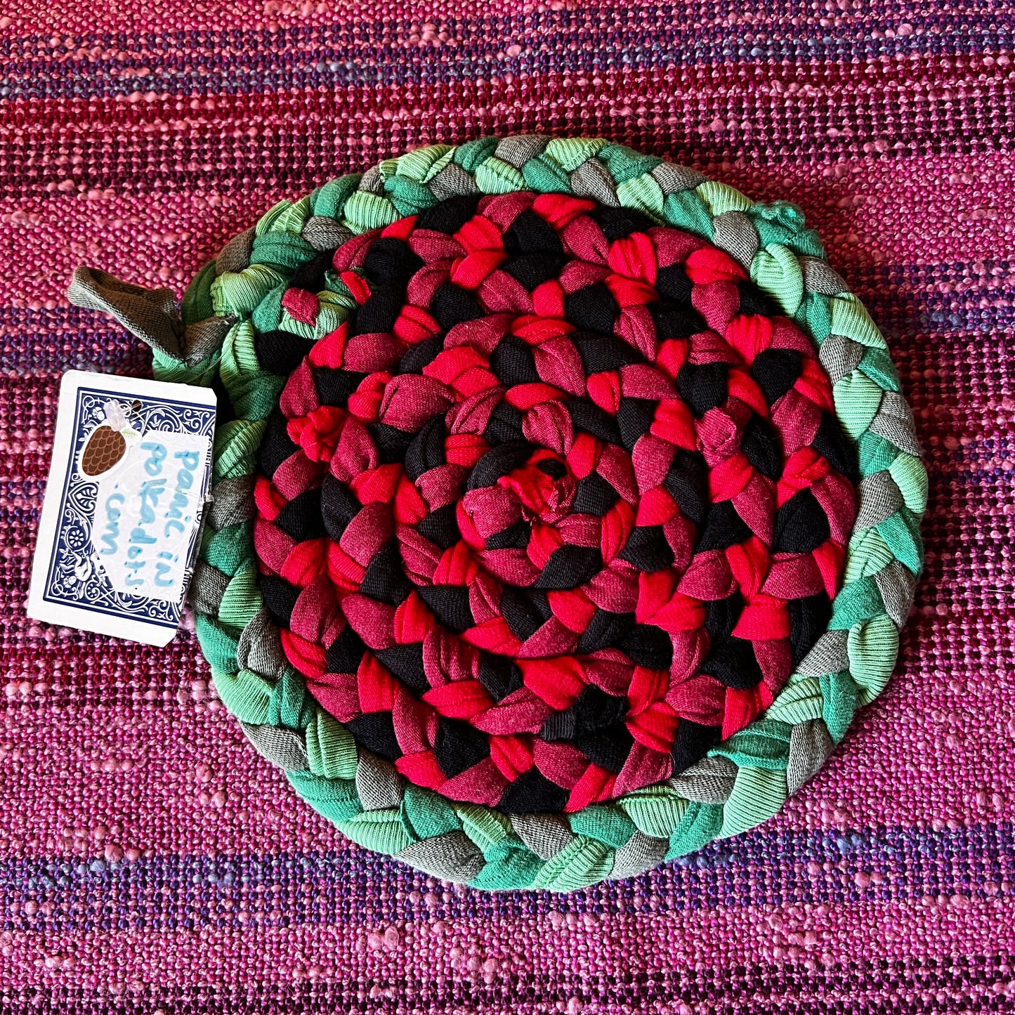 single hand braided and hand sewn potholder/trivet lay flat against a bright pink background, red watermelon