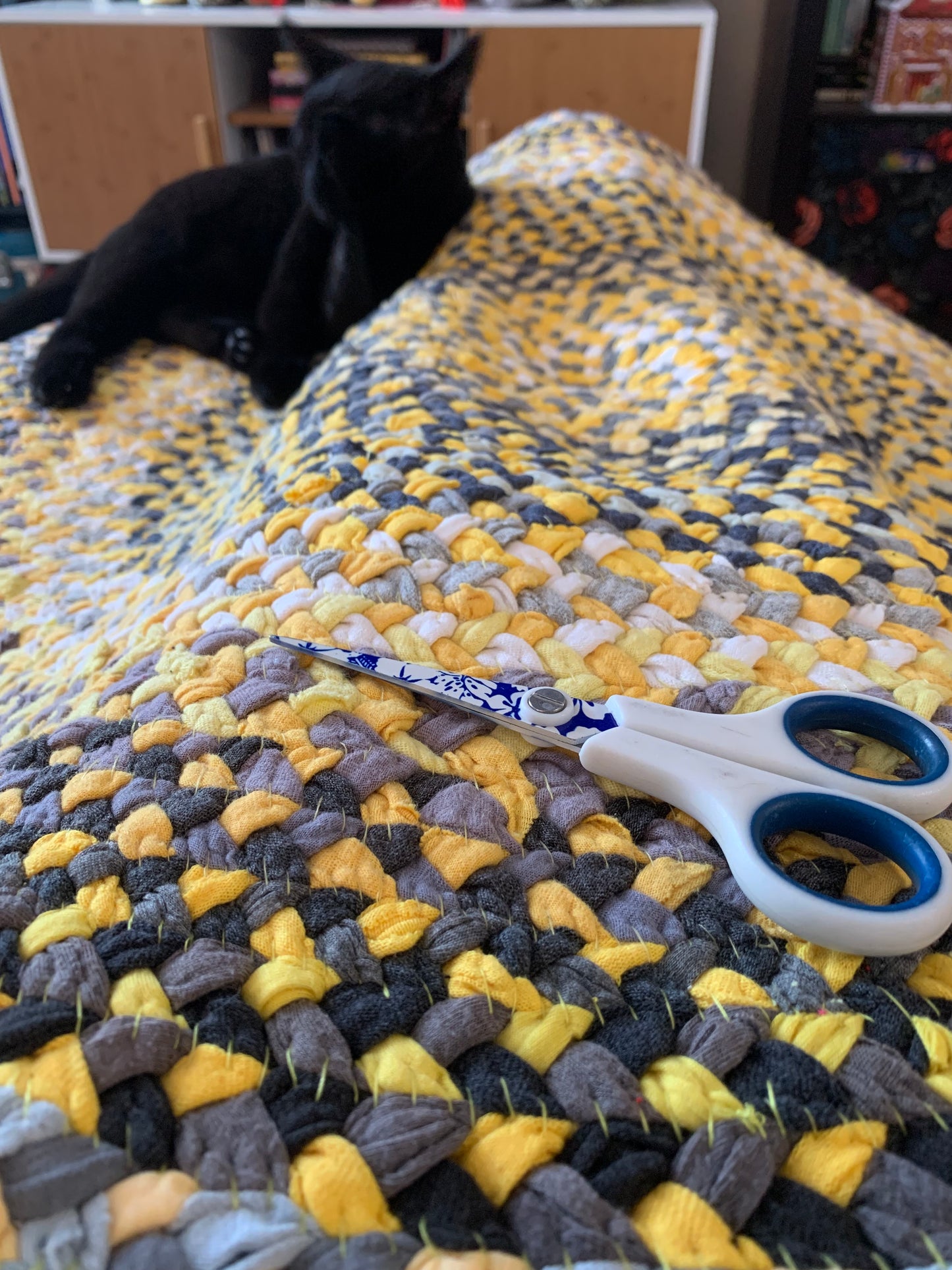 a black cat lays right in the midst of a yellow tshirt rug in progress, scissors and hand-stitches in foreground