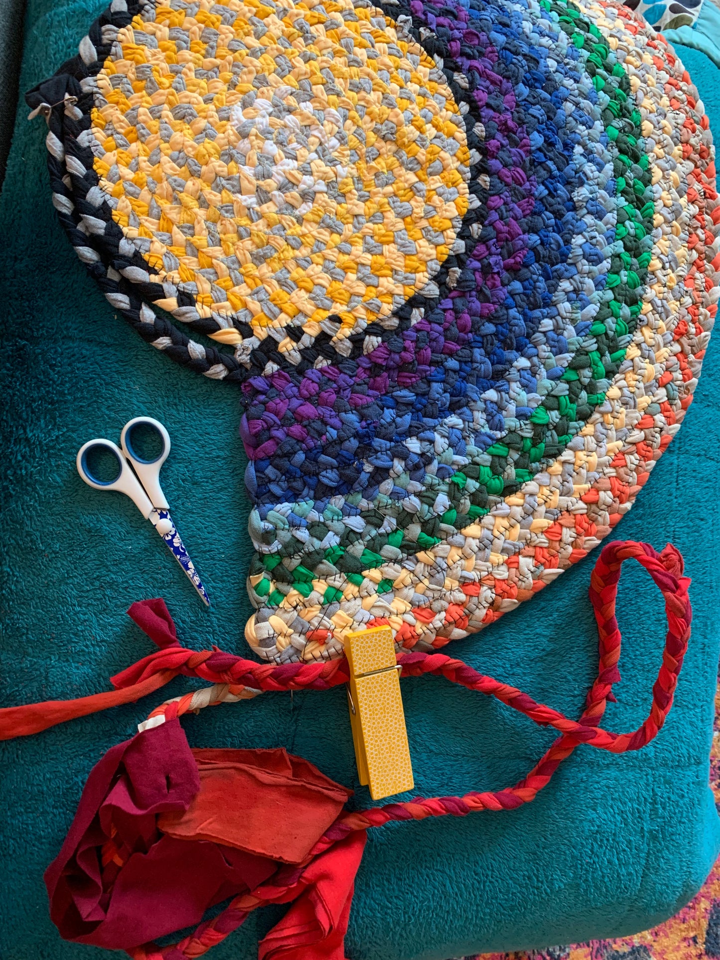 A rainbow rug in progress, on top of a fuzzy blanket, with loose braided red strand and a pair of scissors on top.
