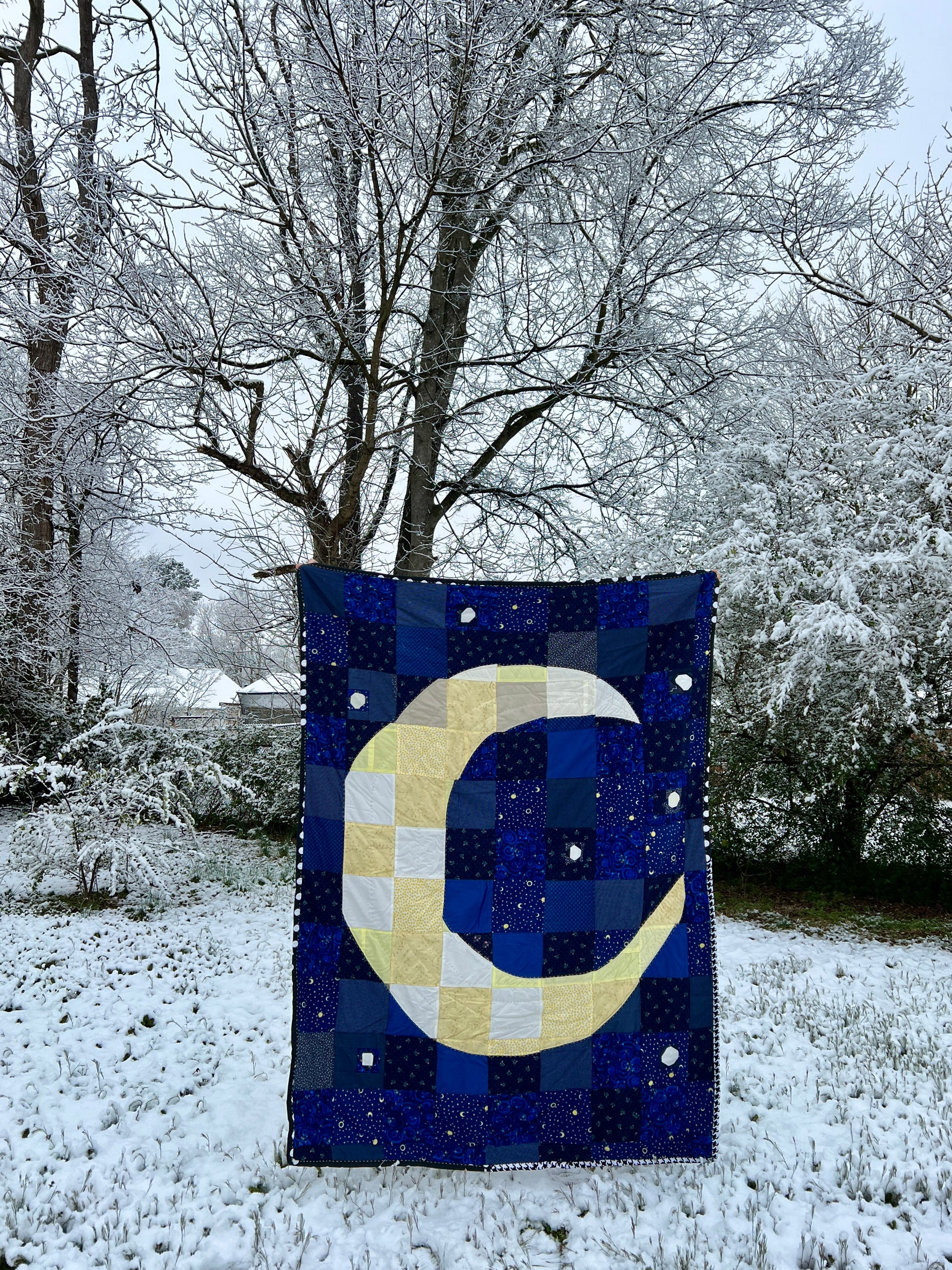 crescent moon quilt, held up outside with snowy trees and snow on the ground in winter