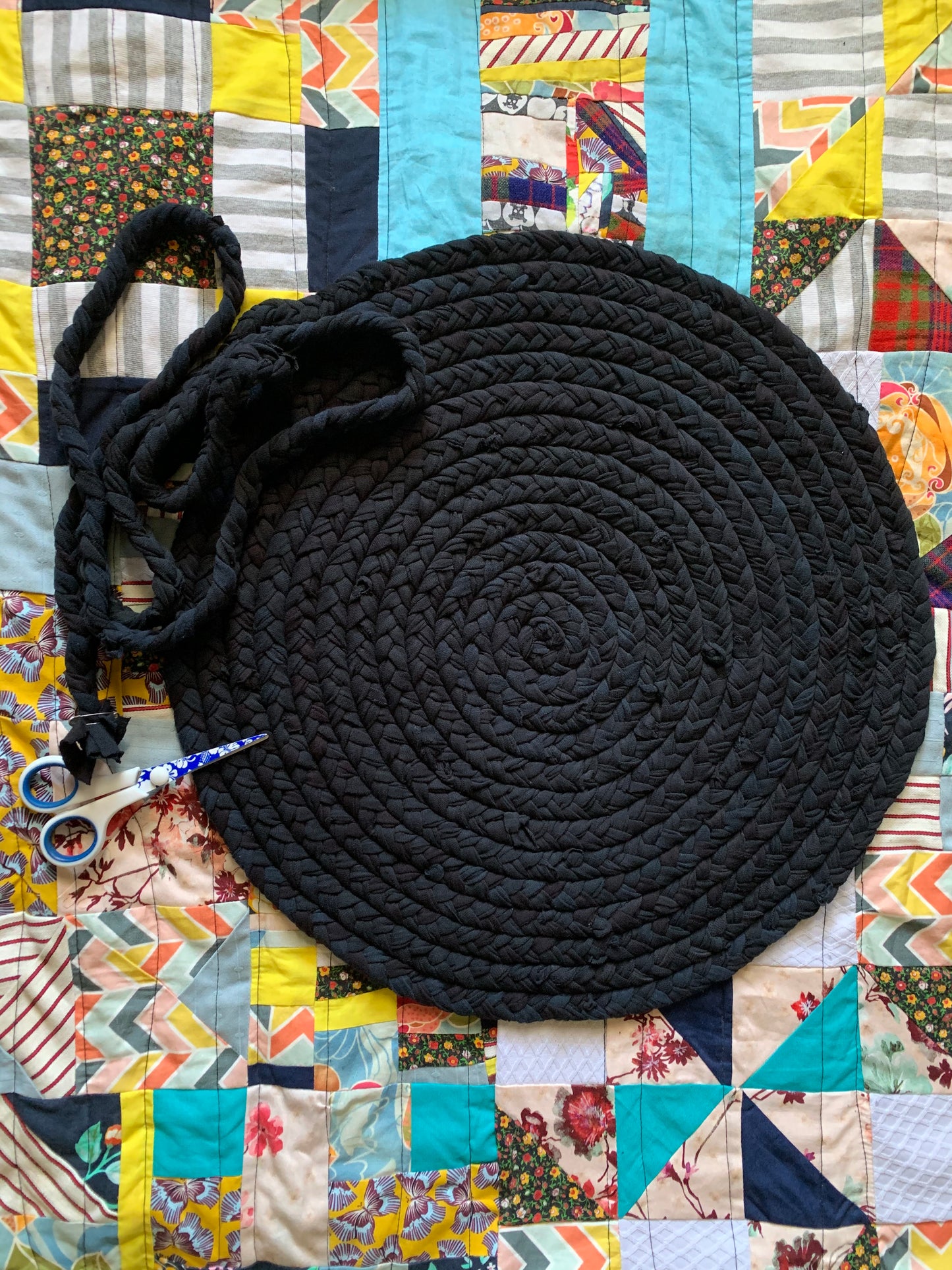 All Black tshirt rug, in progress, partial braid awaits being hand-sewn onto the rest of rug, and a pair of scissors, all flat lay on top of a colorful quilt