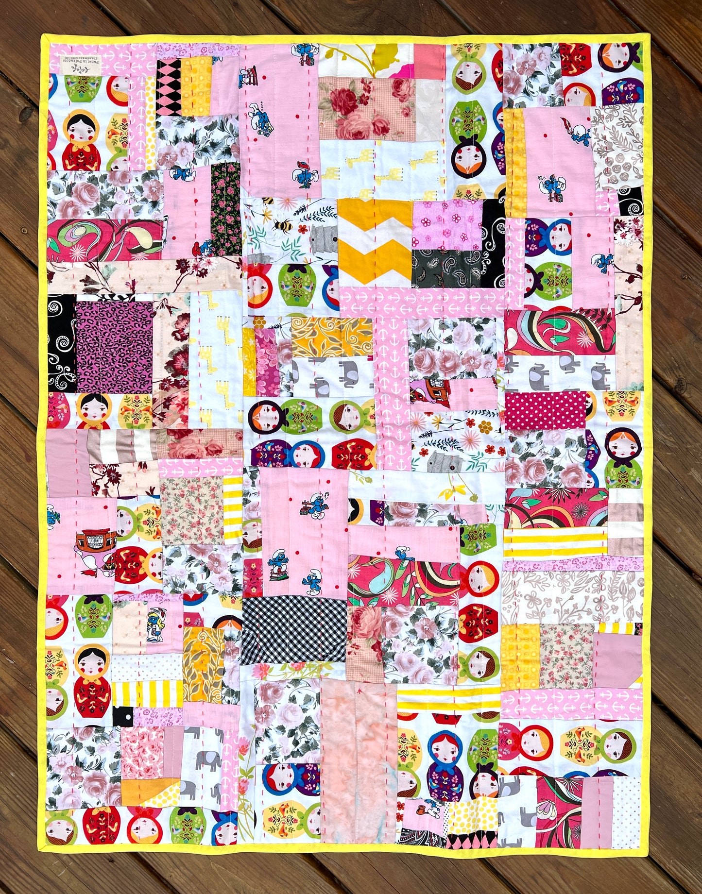 A very vibrant yellow and pink scrappy quilt, outside against a wood deck background