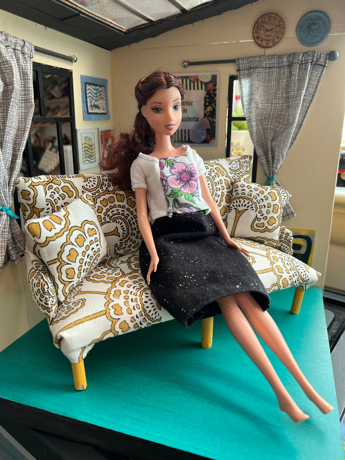 A barbie doll sits on a handmade doll couch, within a dollhouse setting.