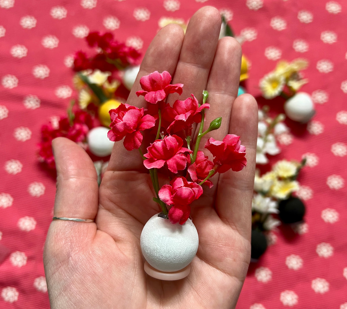 Hand holding a small white vase with red flowers against a pink polka dot background