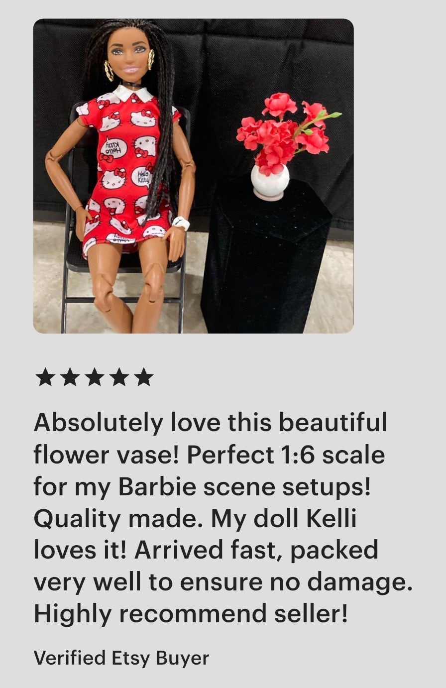 Barbie doll wearing a red dress with white flowers, standing next to a black suit jacket with a flower vase on a gray background.