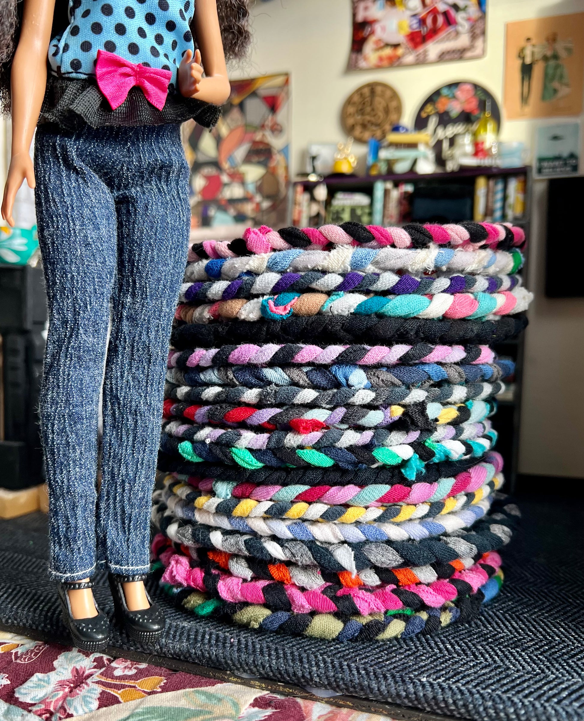 Stack of colorful braided rugs with a doll wearing jeans and a polka dot top in a room setting.