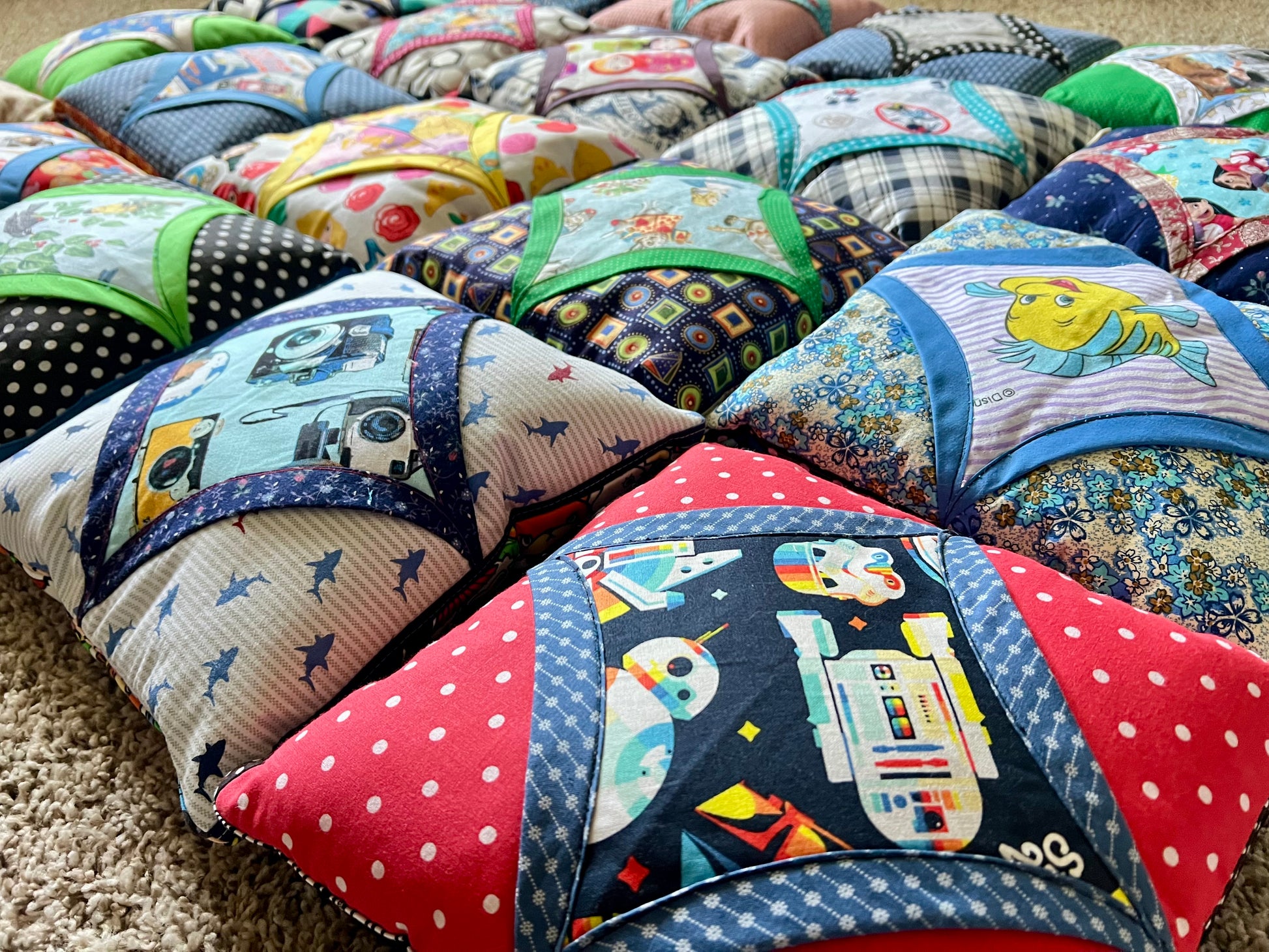 A close-up of a quilted fabric baby support pillow or cushion with a Star Wars R2-D2 design, featuring red polka-dot borders and a navy blue center panel with colorful space-themed graphics. Multiple similar patterned cushions are arranged in rows on a beige carpet.