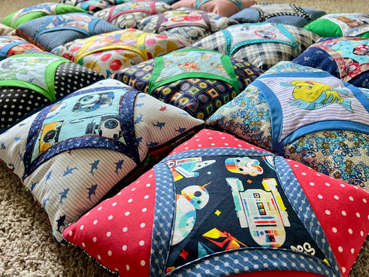 A close-up of a quilted fabric baby support pillow or cushion with a Star Wars R2-D2 design, featuring red polka-dot borders and a navy blue center panel with colorful space-themed graphics. Multiple similar patterned cushions are arranged in rows on a beige carpet.
