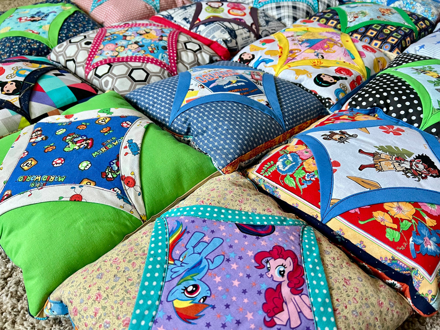 A collection of colorful, handmade quilted pillows featuring various cartoon and animal prints, arranged closely together on a carpeted floor.