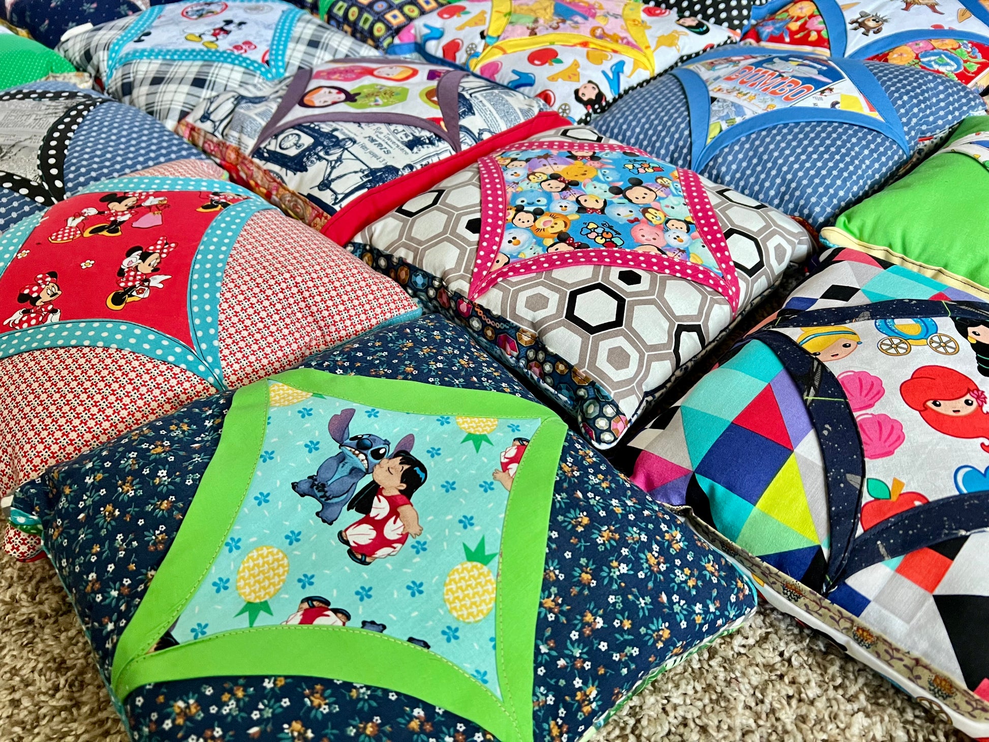 A close-up of a colorful patchwork quilted cushion or pillow set with various cartoon and geometric patterns, including various characters and abstract shapes, arranged on a beige carpet.