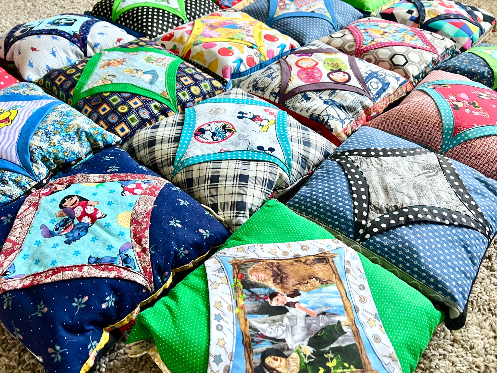 A collection of handmade quilted baby pillows arranged together on a beige carpet. The pillows feature a variety of colorful, patterned fabrics including plaid, polka dots, and character-themed prints.
