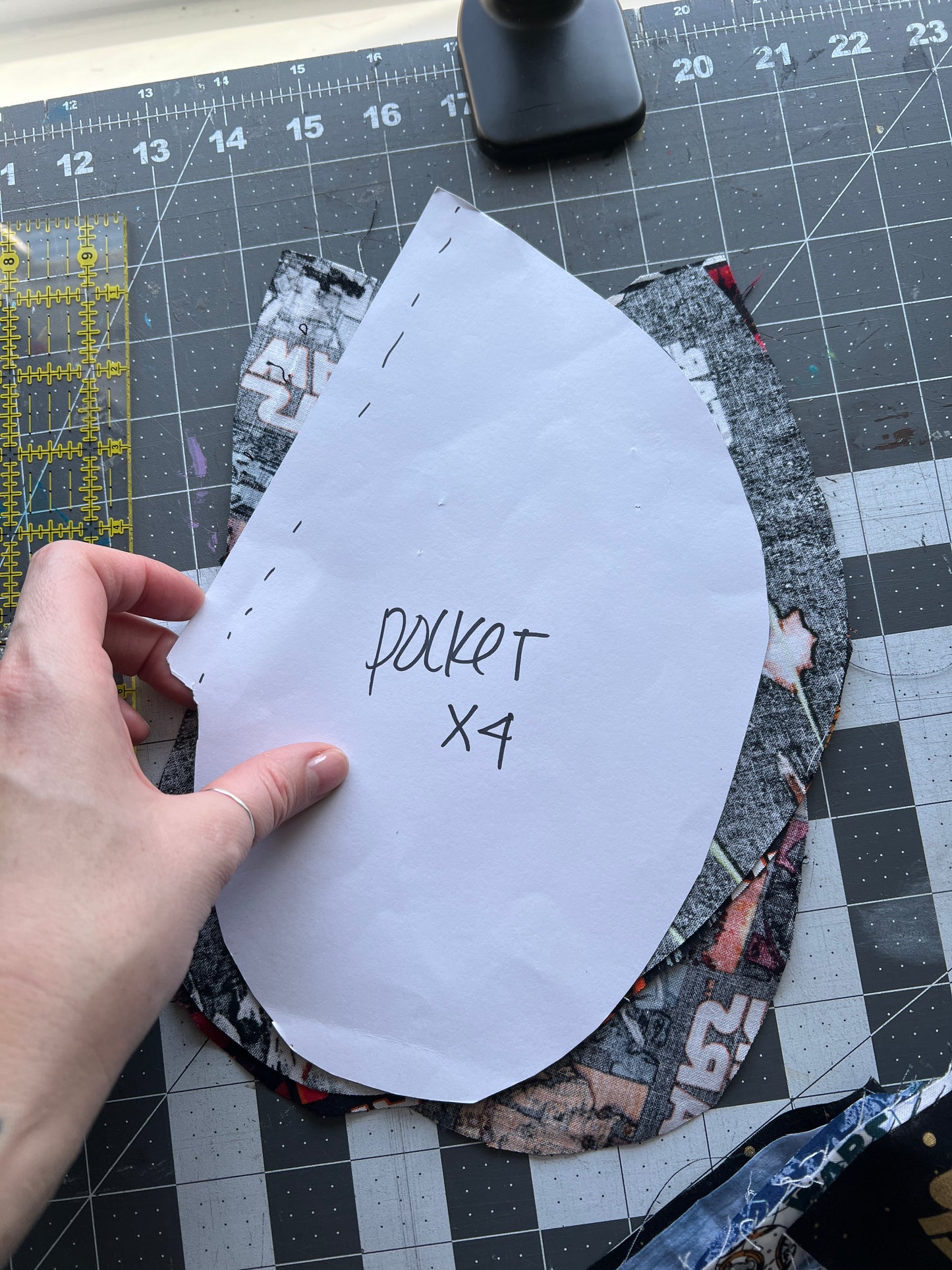 Hand holding a paper pattern labeled 'pocket X4' on a cutting mat with fabric pieces.