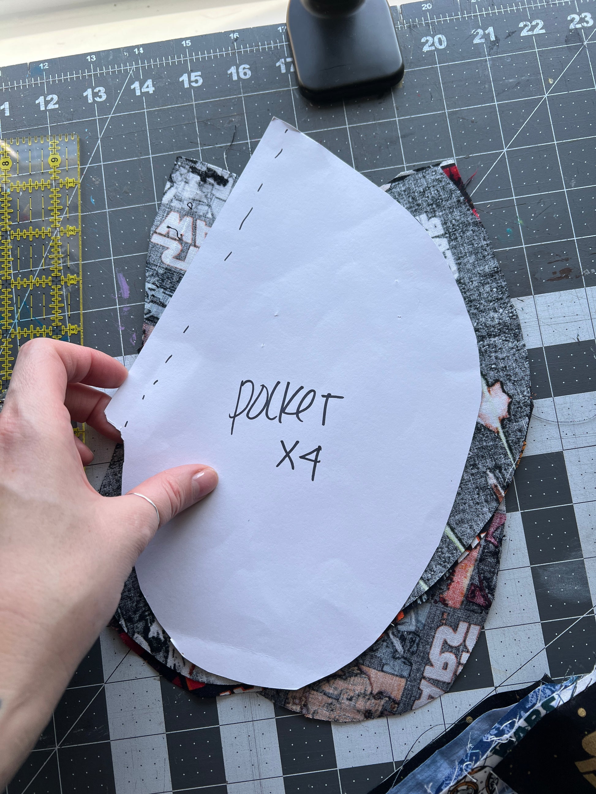 Hand holding a paper pattern labeled 'pocket X4' on a cutting mat with fabric pieces.