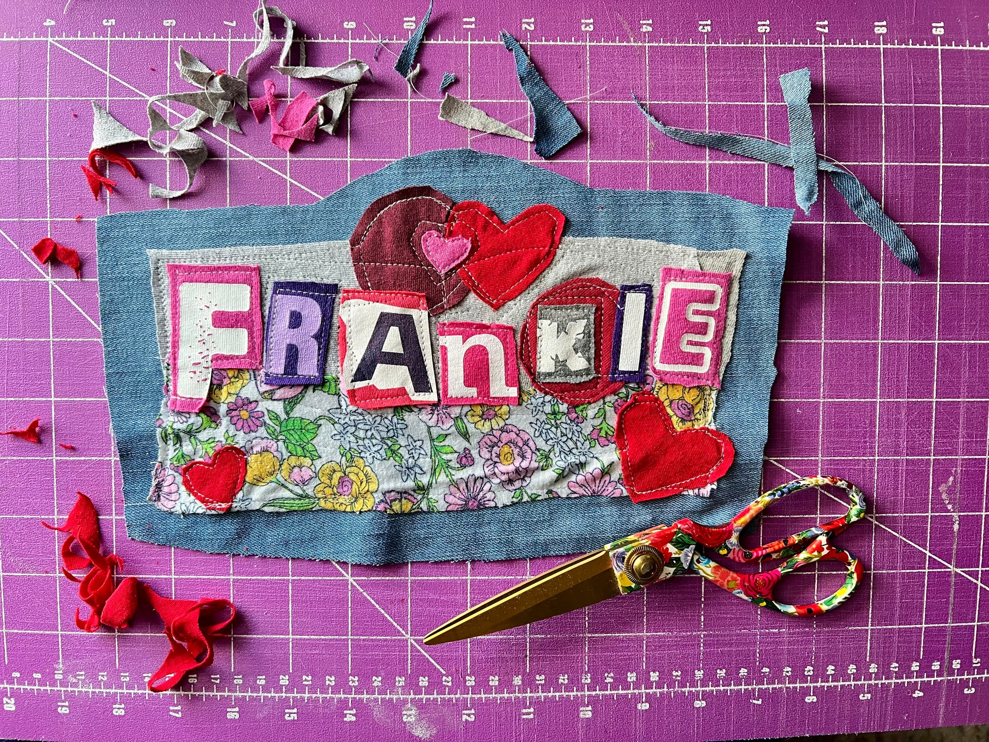 A handmade fabric patch with the name 'FRANKIE' spelled out in colorful letters, decorated with red hearts and a floral pattern, placed on a purple cutting mat with scissors and fabric scraps around it.