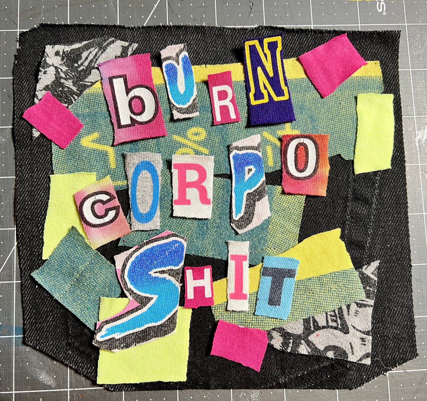 A square handmade patch featuring a colorful collage of cut paper and fabric pieces spelling out 'BURN CORPO SHIT' in bold letters, set against a dark denim-like background on a gray cutting mat.