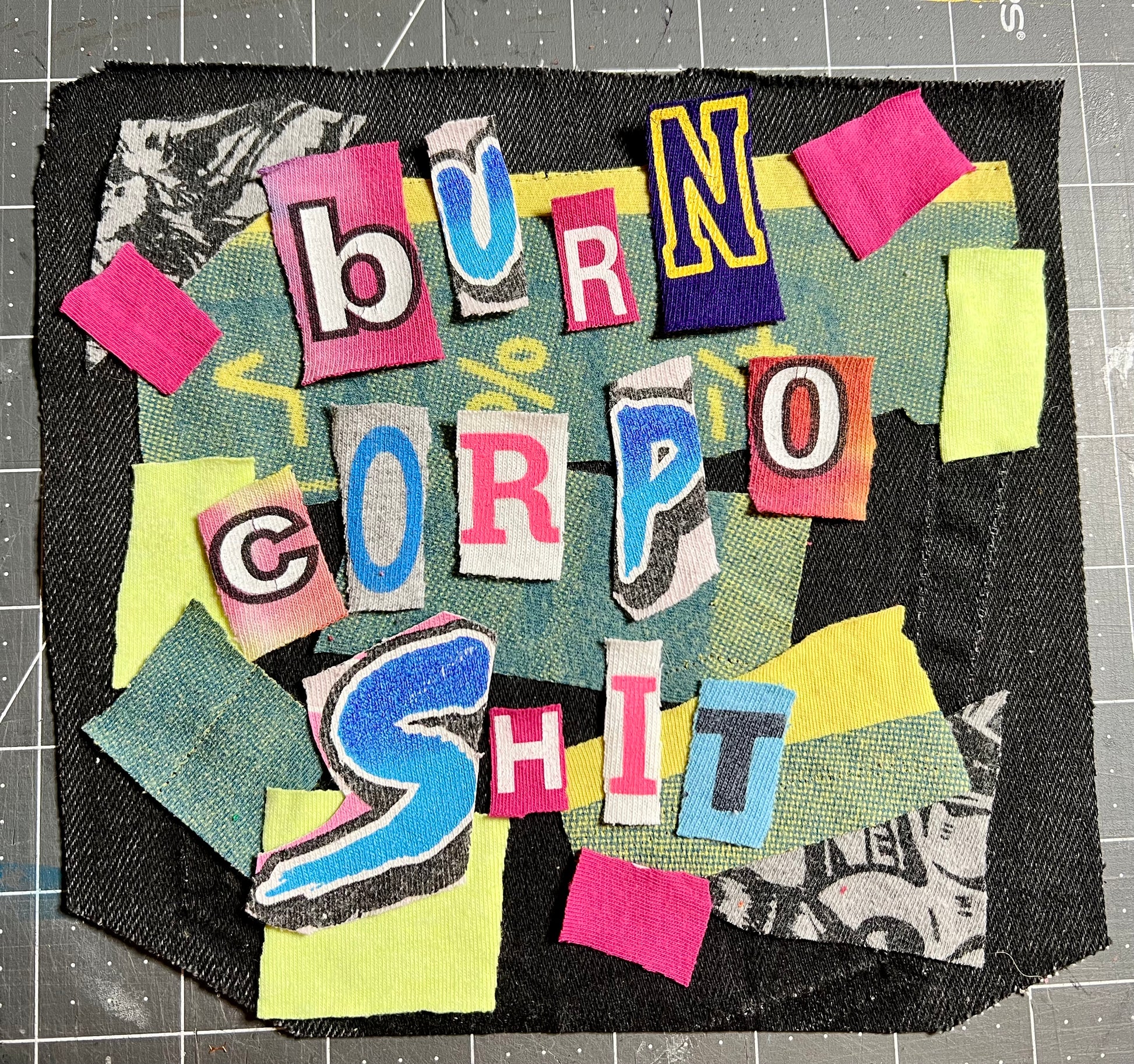 A square handmade patch featuring a colorful collage of cut paper and fabric pieces spelling out 'BURN CORPO SHIT' in bold letters, set against a dark denim-like background on a gray cutting mat.