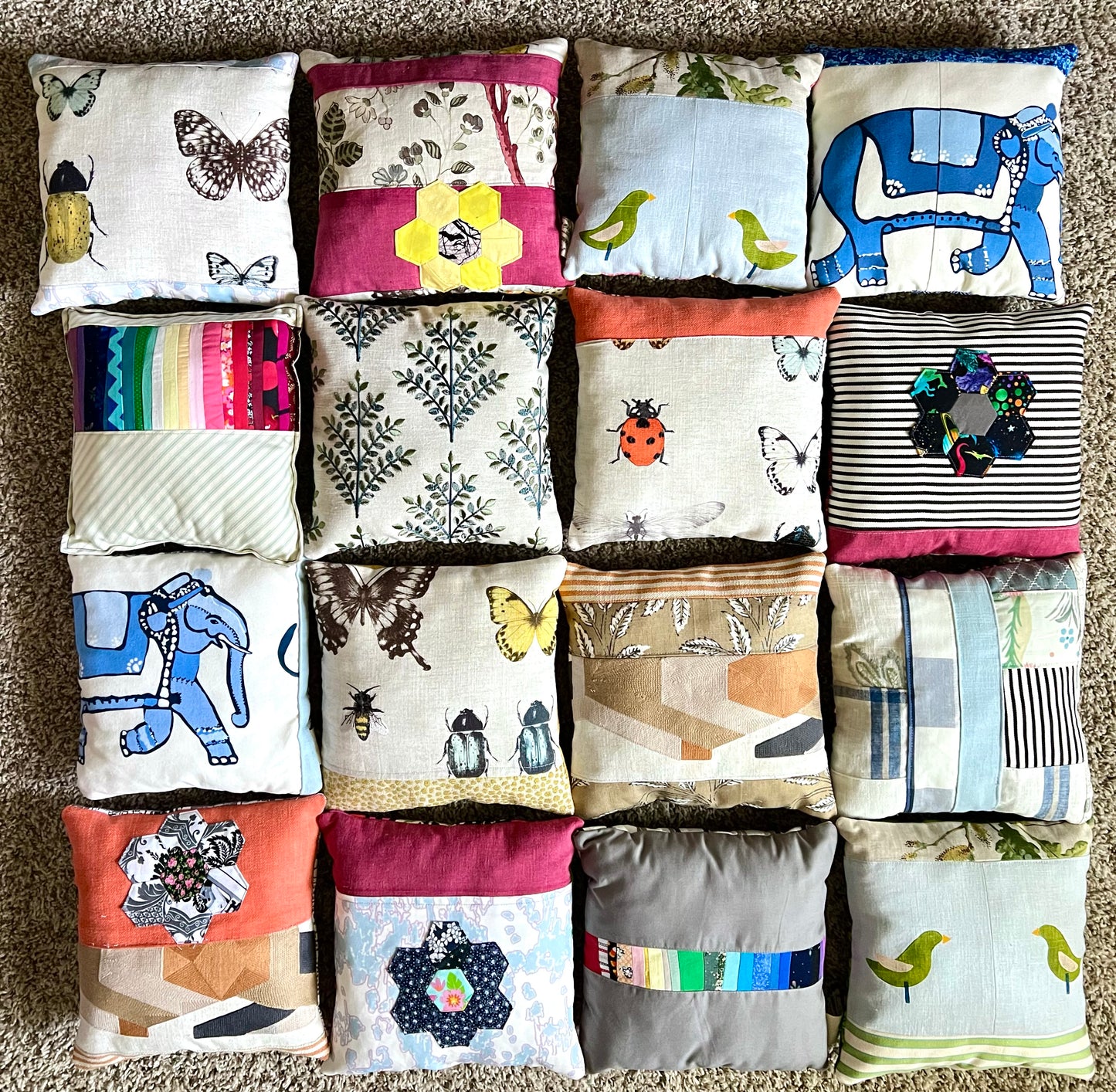 A 4x4 grid of small square fabric pillows laid flat on a beige carpet, each with different printed or sewn designs including butterflies, birds, elephants, stripes, flowers, and geometric patterns.